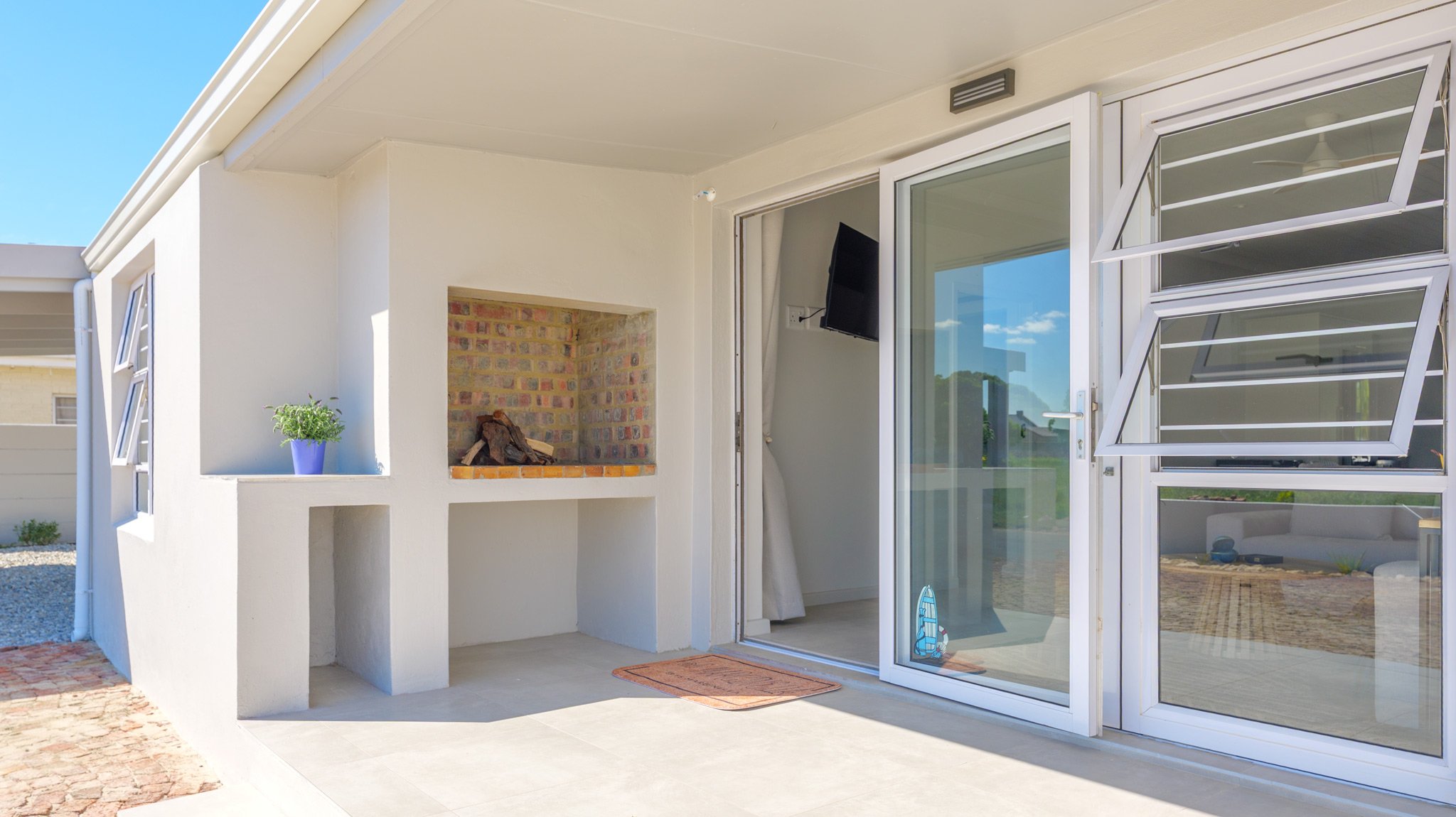 Patio area featuring a built-in brick fireplace with wood, a small potted plant in a purple pot, and a sliding glass door with a TV mounted inside. There are open exterior windows with louvers, and a rubber doormat in front of the door. The scene is 