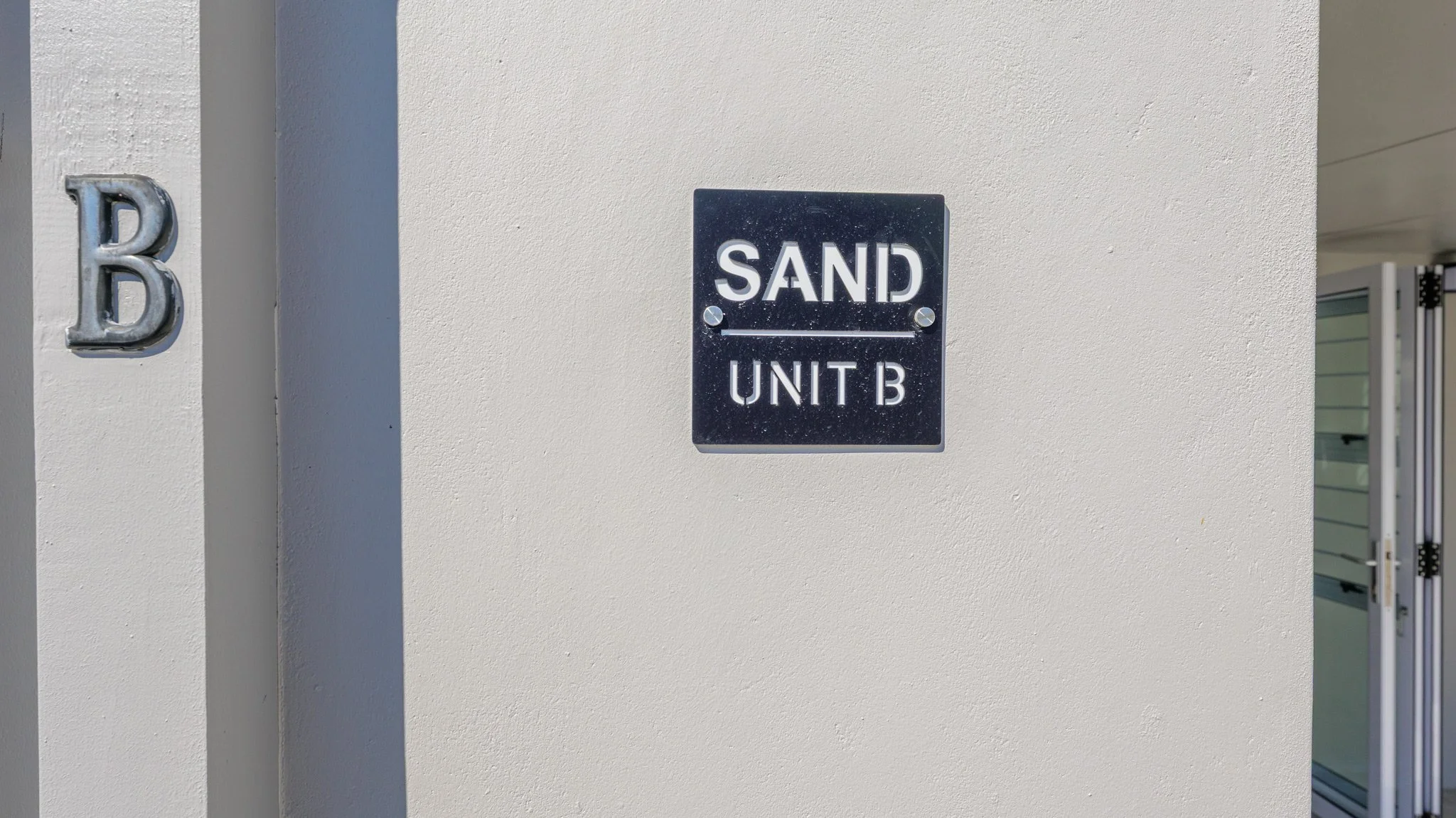 Close-up photo of a black metal sign on a white wall indicating 'SAND UNIT B' with an embossed metal letter 'B' on the adjacent wall.