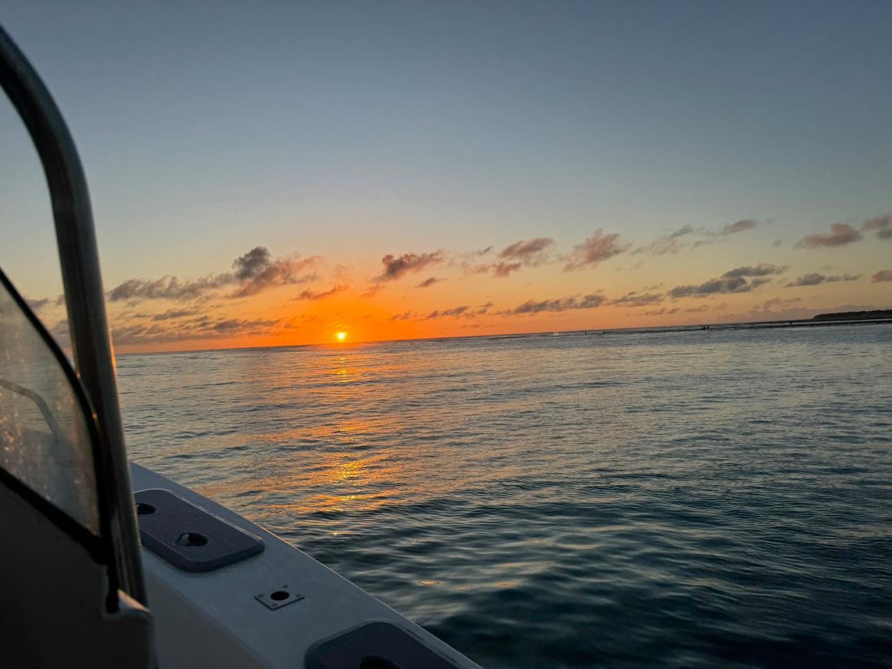Sunset over the ocean as seen from a boat, with a part of the boat's railing and deck visible in the foreground.