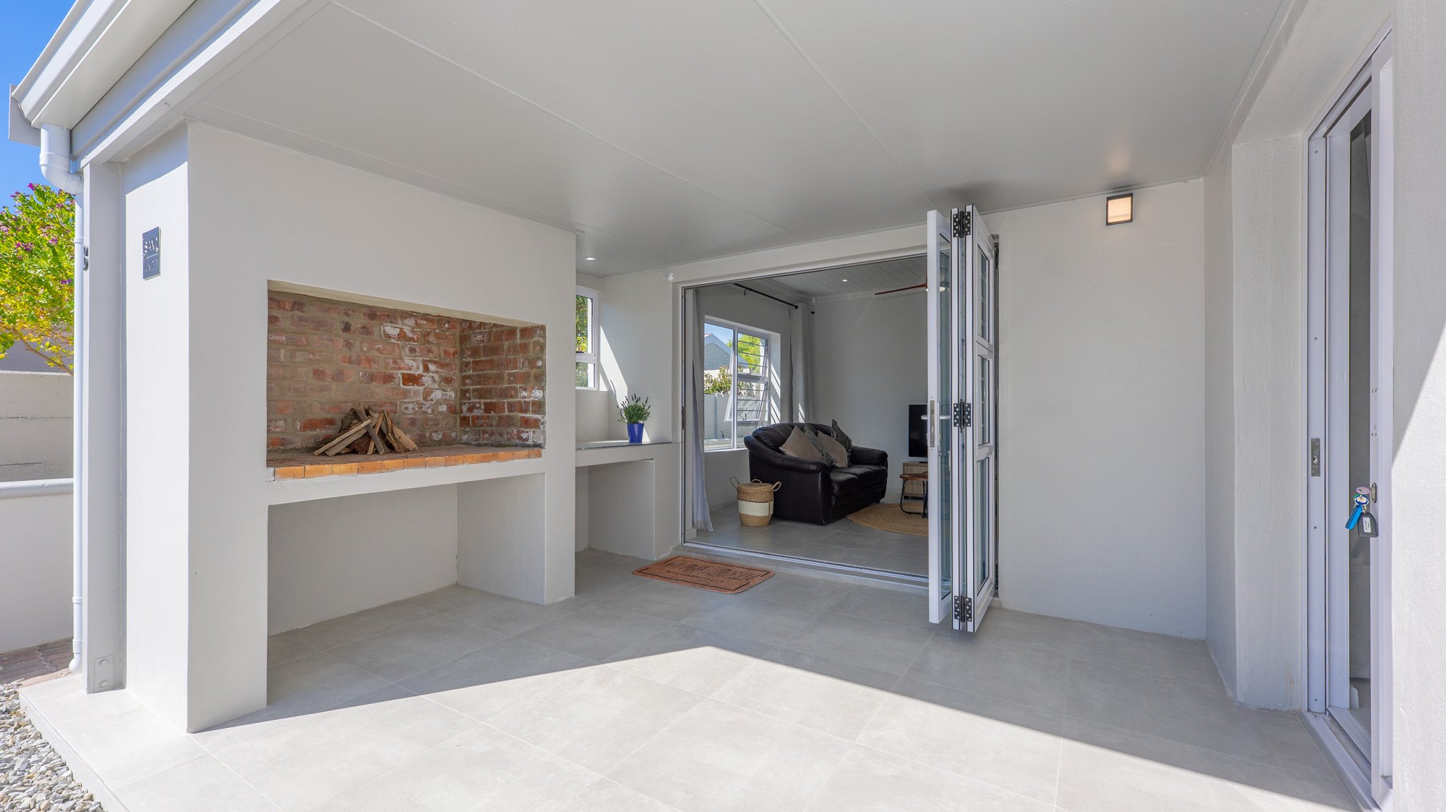 Outdoor patio area with brick fireplace, gray tiled floor, and open glass door leading to living room with black sofa and large windows.