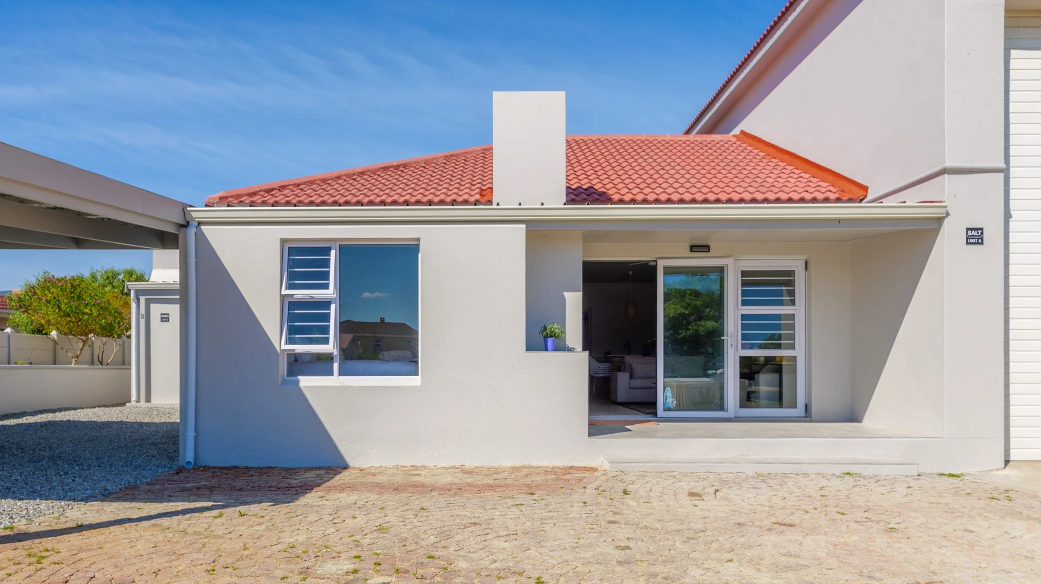 Modern house with white exterior walls, red tiled roof, sliding glass door, and a small porch with a potted plant, situated under a bright blue sky with a few trees in background.