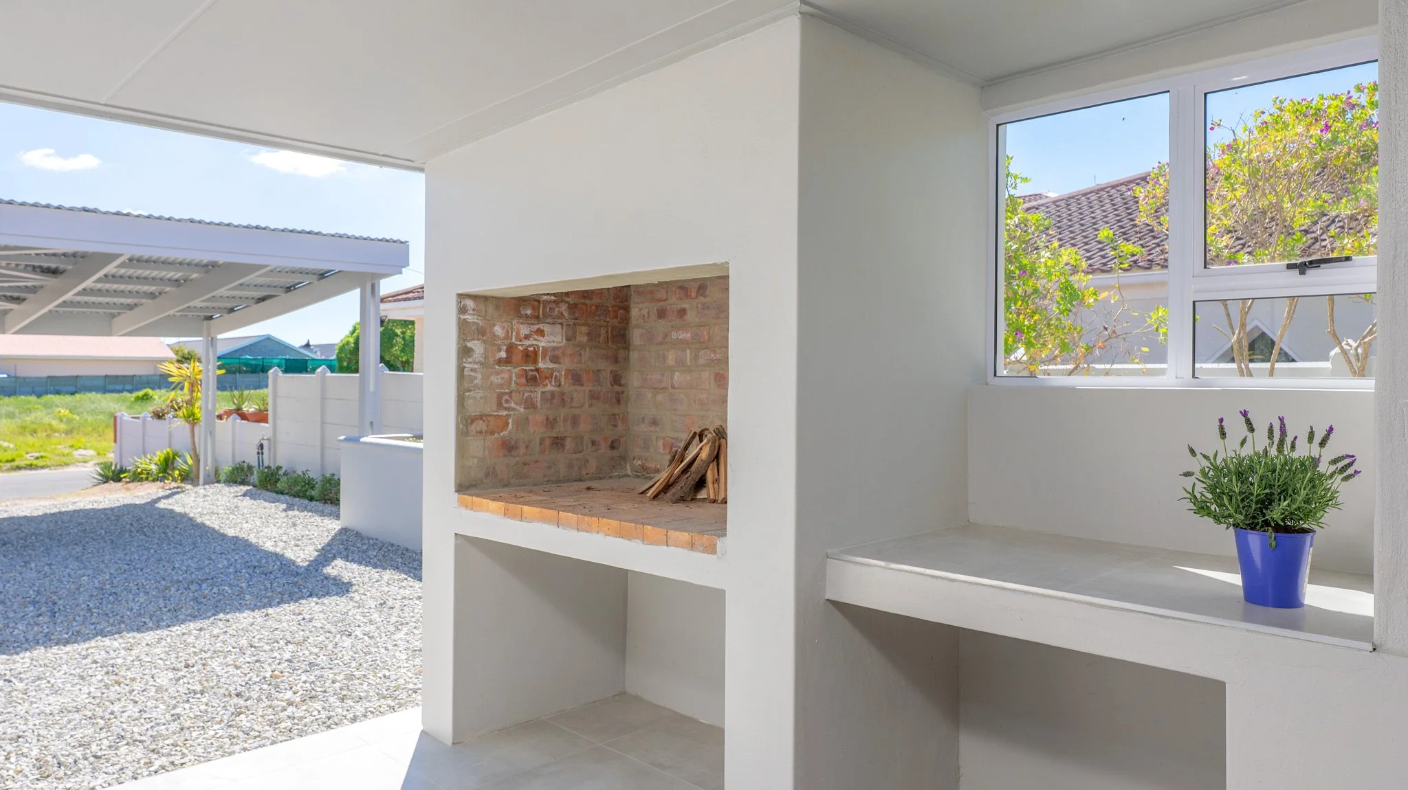 Outdoor patio with brick fireplace, white walls, window with view of trees, potted lavender plant, gravel ground, and a covered carport.
