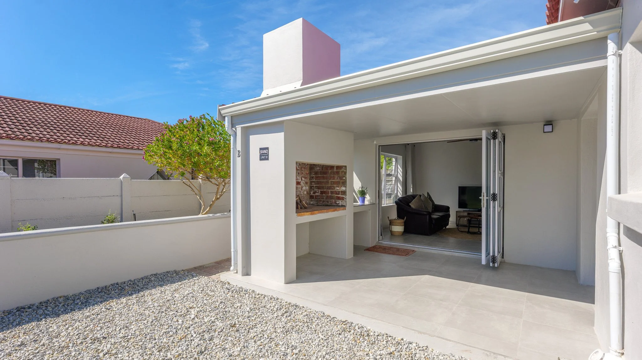 View of a backyard with an outdoor patio area, showing gravel ground, a small tree, and a white building with a sliding glass door leading to a living room with a television and black sofa inside.