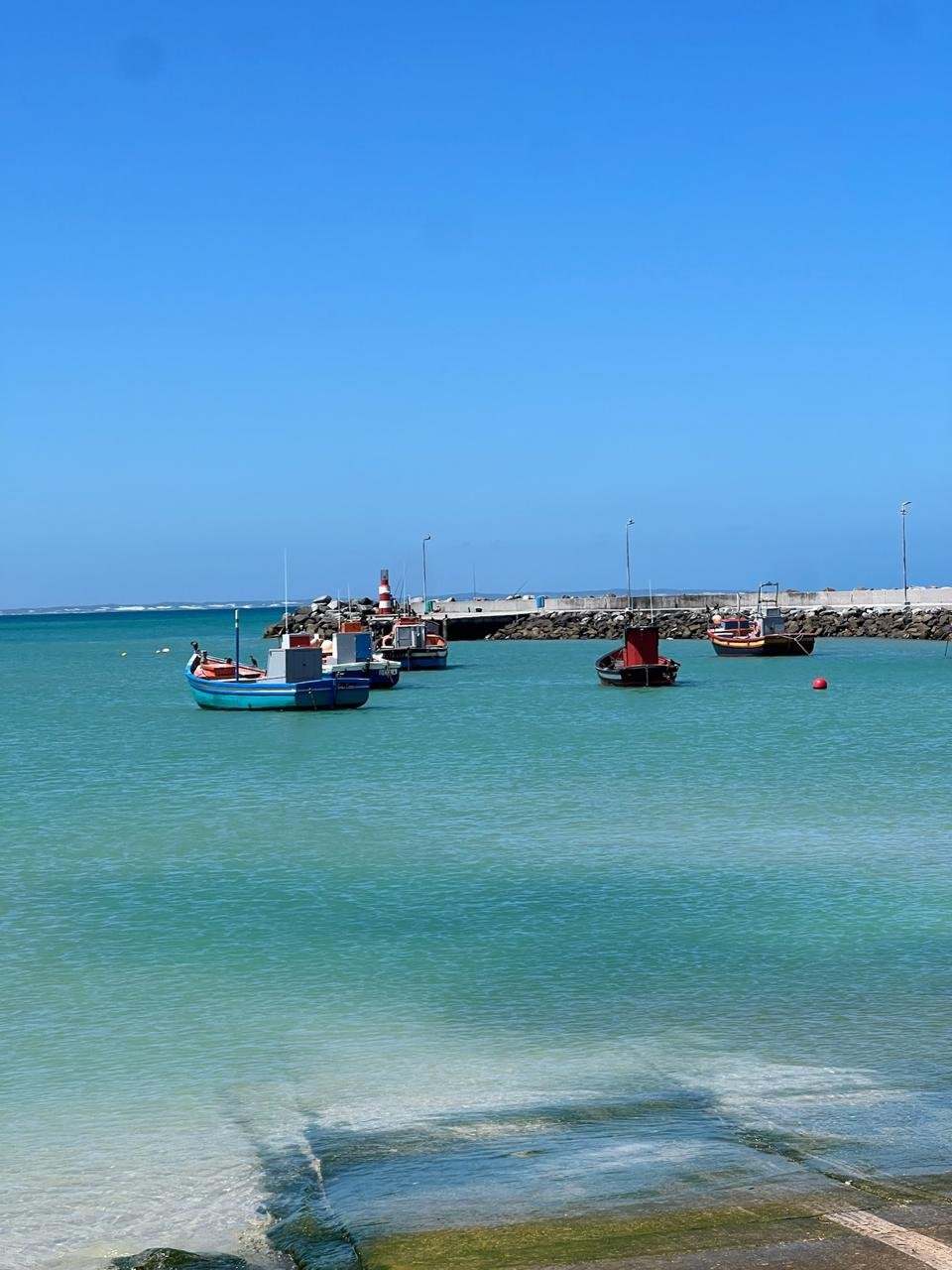 Boats docked in a turquoise harbor near a concrete pier with a lighthouse and lined with streetlights, under a clear blue sky.