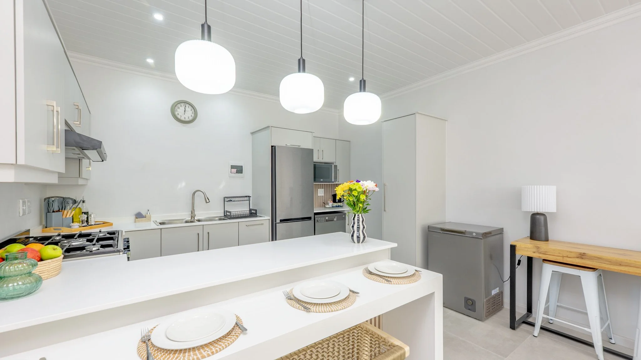 Modern kitchen with white cabinets, stainless steel refrigerator, microwave, and a white island counter with three place settings and a vase of colorful flowers. There are three hanging white pendant lights, a wall clock, a lamp, and a small wooden t