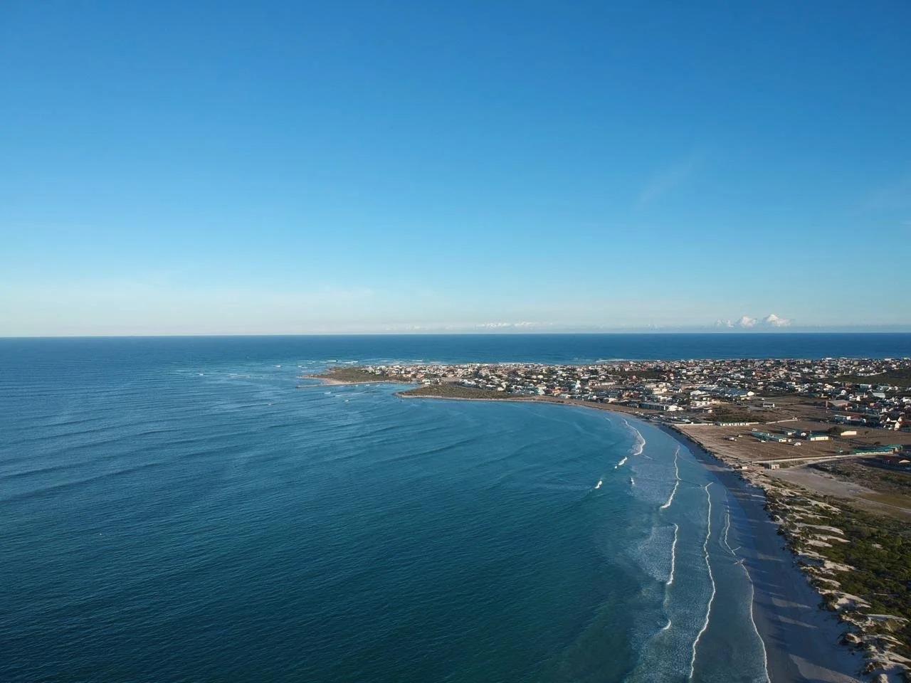 Aerial view of a coastal city with a long shoreline, waves hitting the beach, and a clear blue sky.