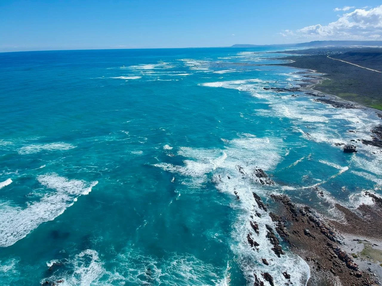 Aerial view of a rugged coastline with blue ocean waves crashing against rocks, extending to distant mountains under a partly cloudy sky