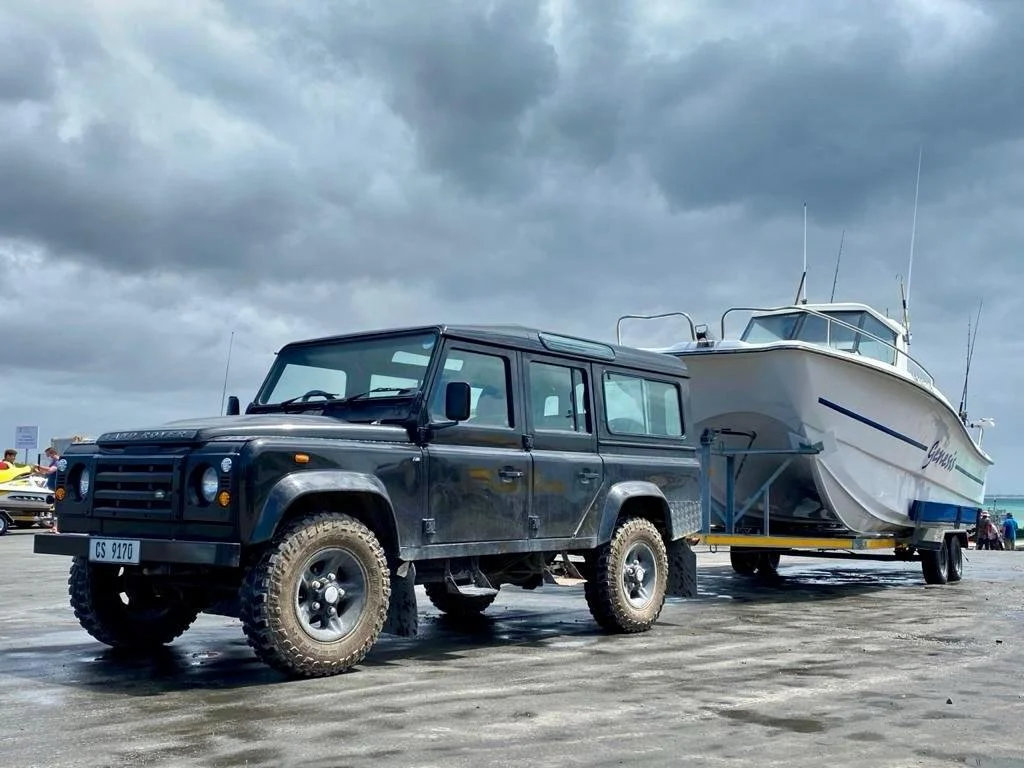 A black Land Rover Defender SUV with mud tires is parked on a wet concrete surface, attached to a trailer carrying a large white boat with a cabin, under a cloudy sky.