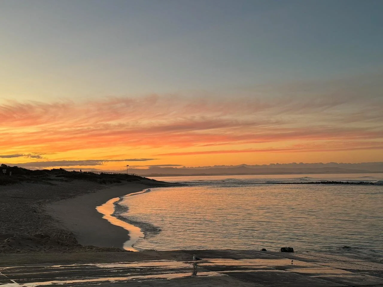 Sunset over a calm beach with a partly cloudy sky, gentle waves lapping the shore, and a wooden pathway leading towards the horizon.