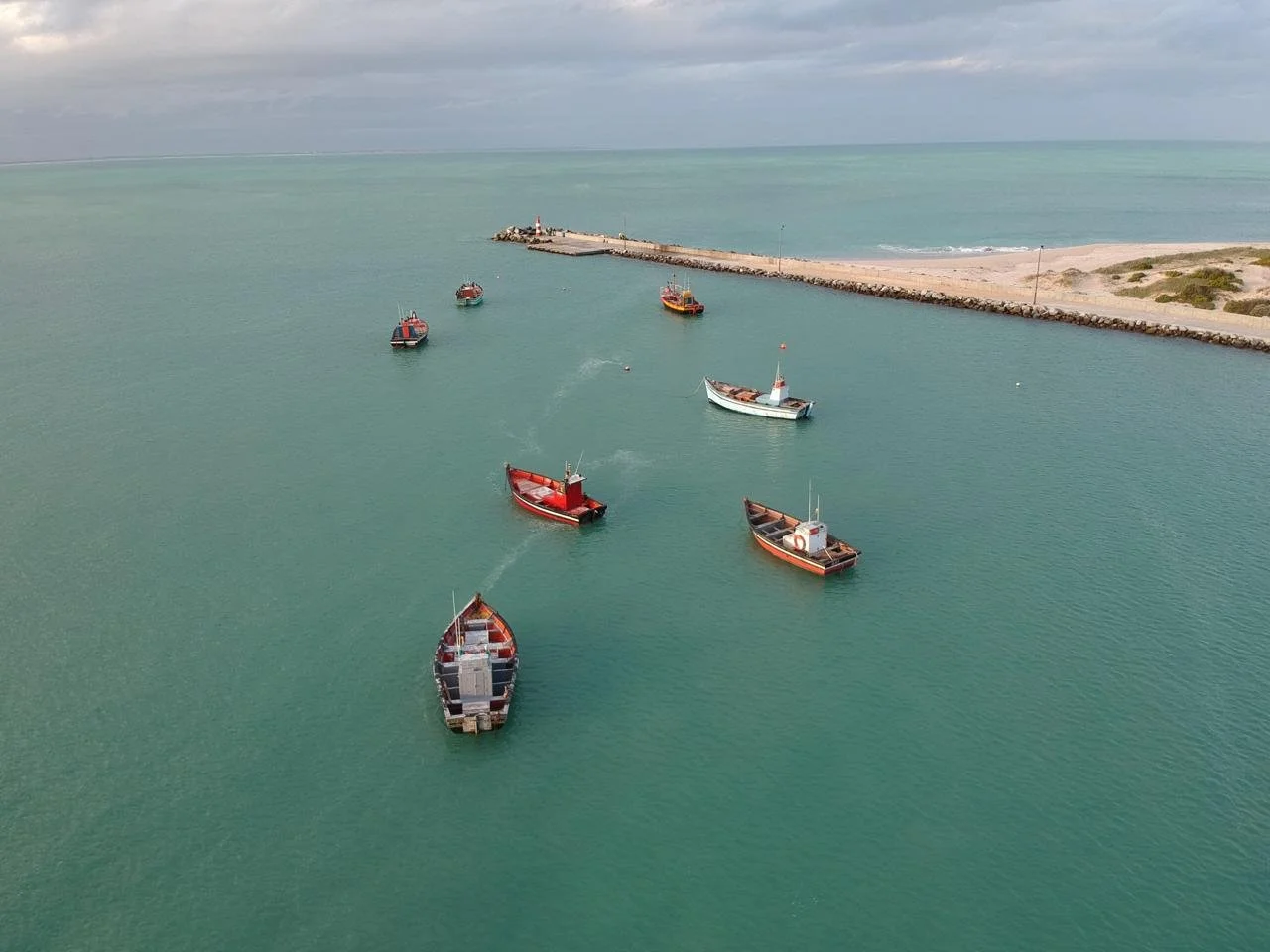 Aerial view of several small boats floating in a harbor near a rocky breakwater and sandy beach under cloudy skies.