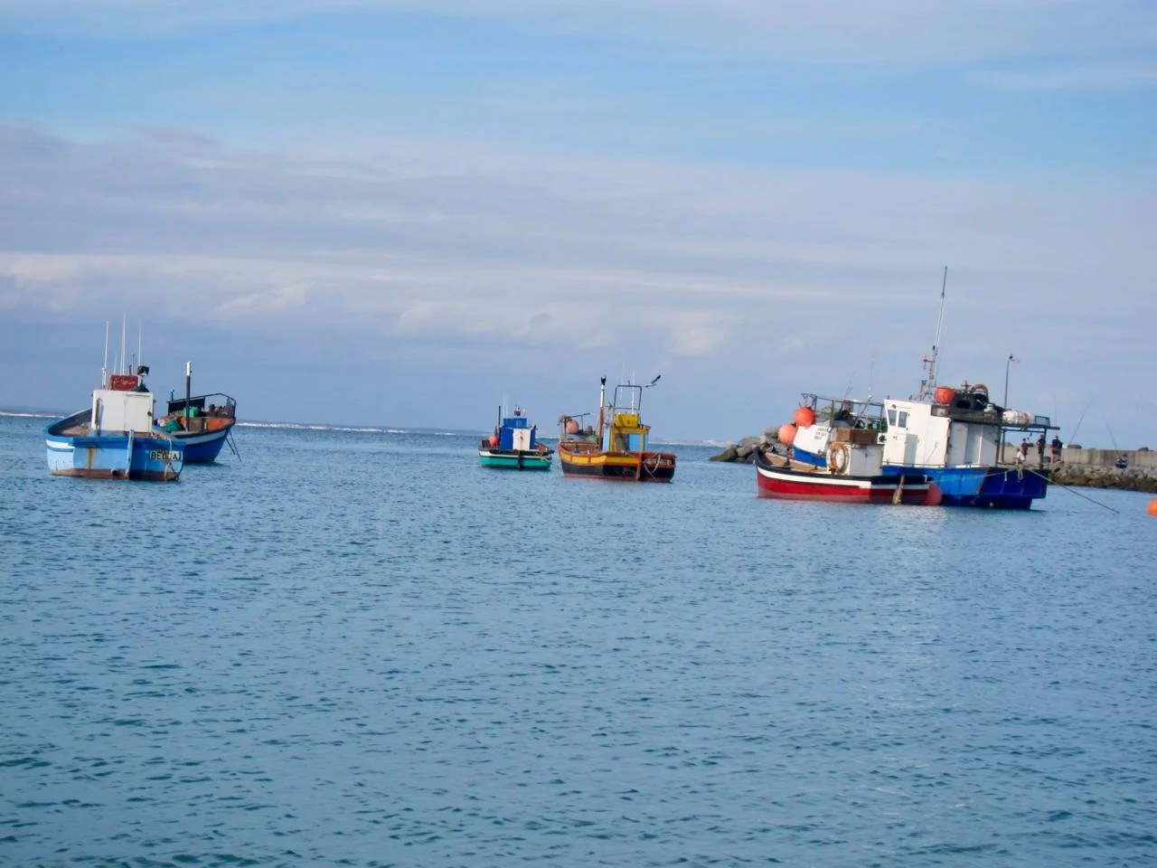 Several colorful boats are anchored in calm water near a rocky breakwater