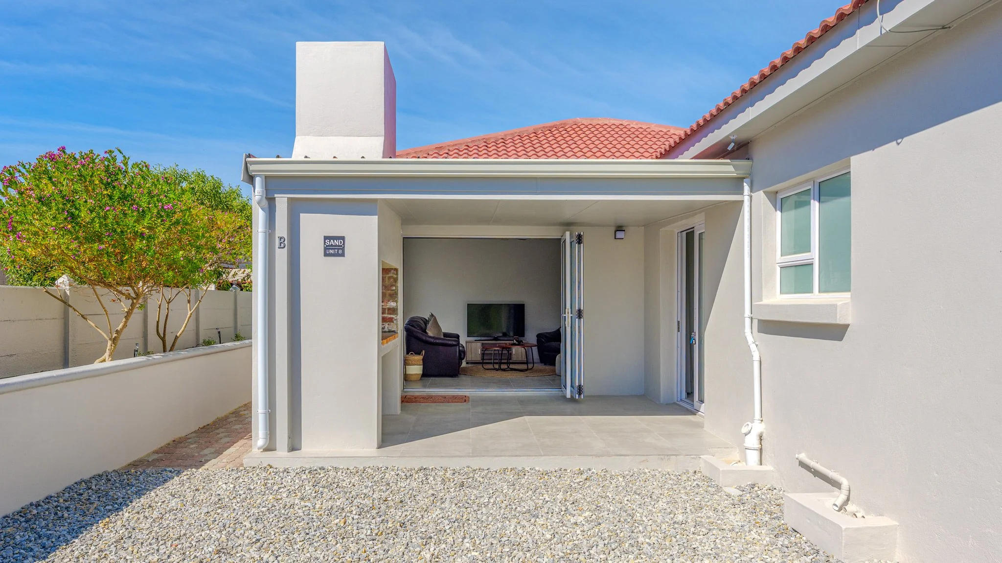 View of a house patio with an open door leading to a living room with a TV, two chairs, and a brick fireplace, surrounded by a white fence, gravel ground, and a tree with pink flowers.