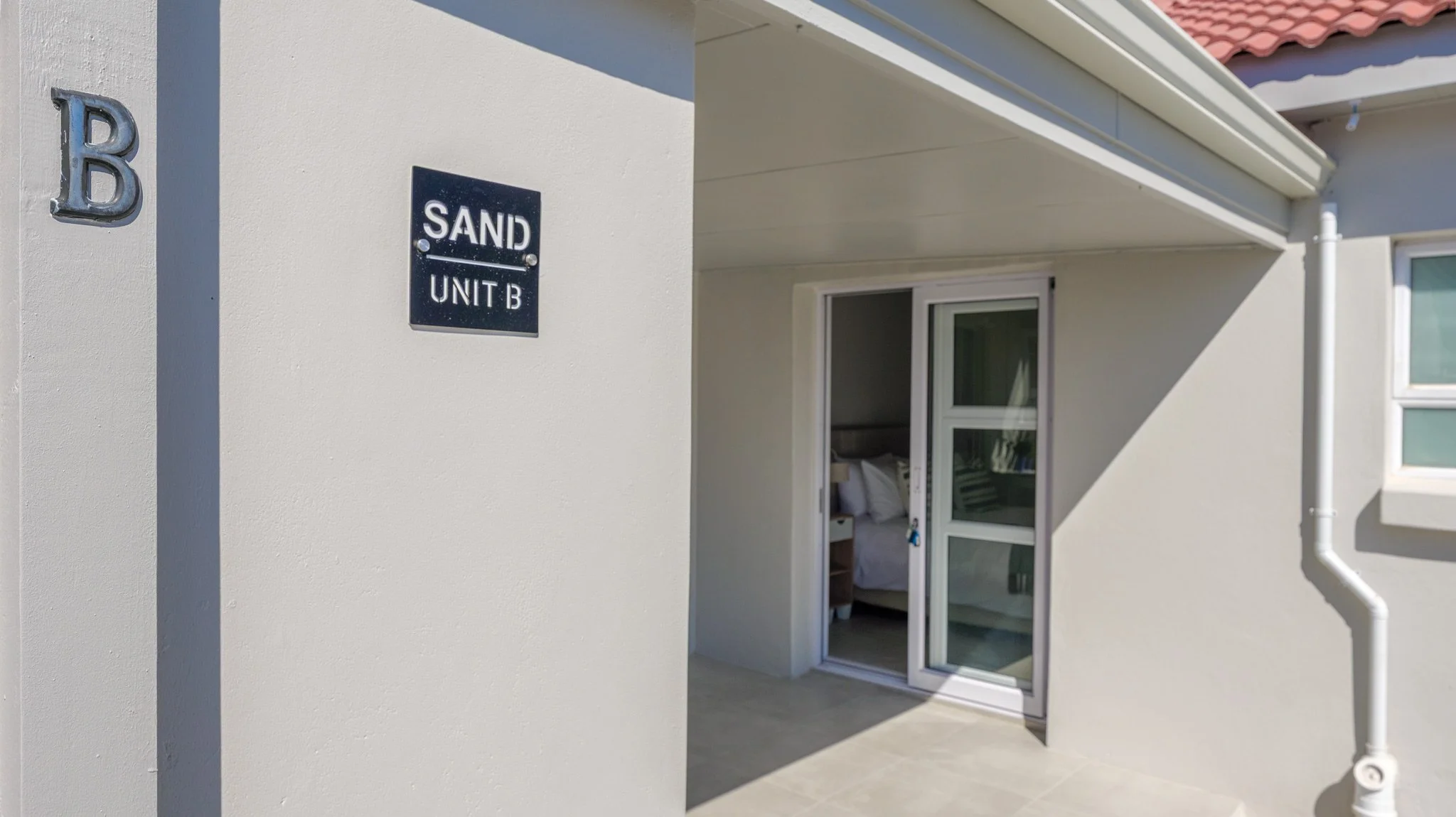 Exterior view of a residence with a sign reading 'Sand Unit B' on a white stucco wall and a sliding glass door leading to a bedroom inside. Part of a red tiled roof is visible.