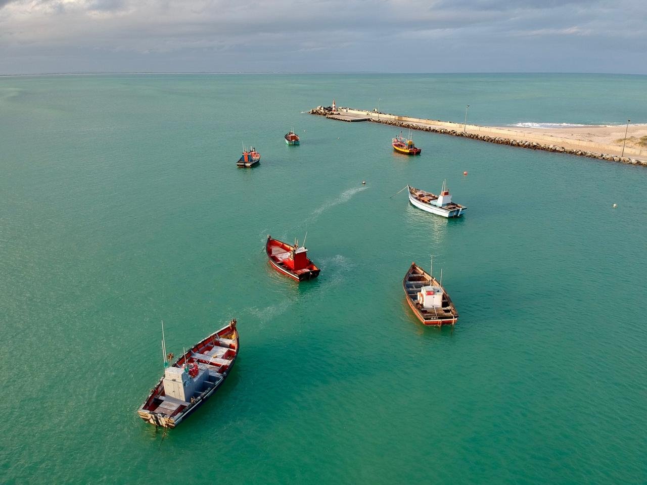 Multiple boats anchored near a breakwater in a calm, greenish-blue harbor with a lighthouse at the end of the breakwater and cloudy sky overhead.