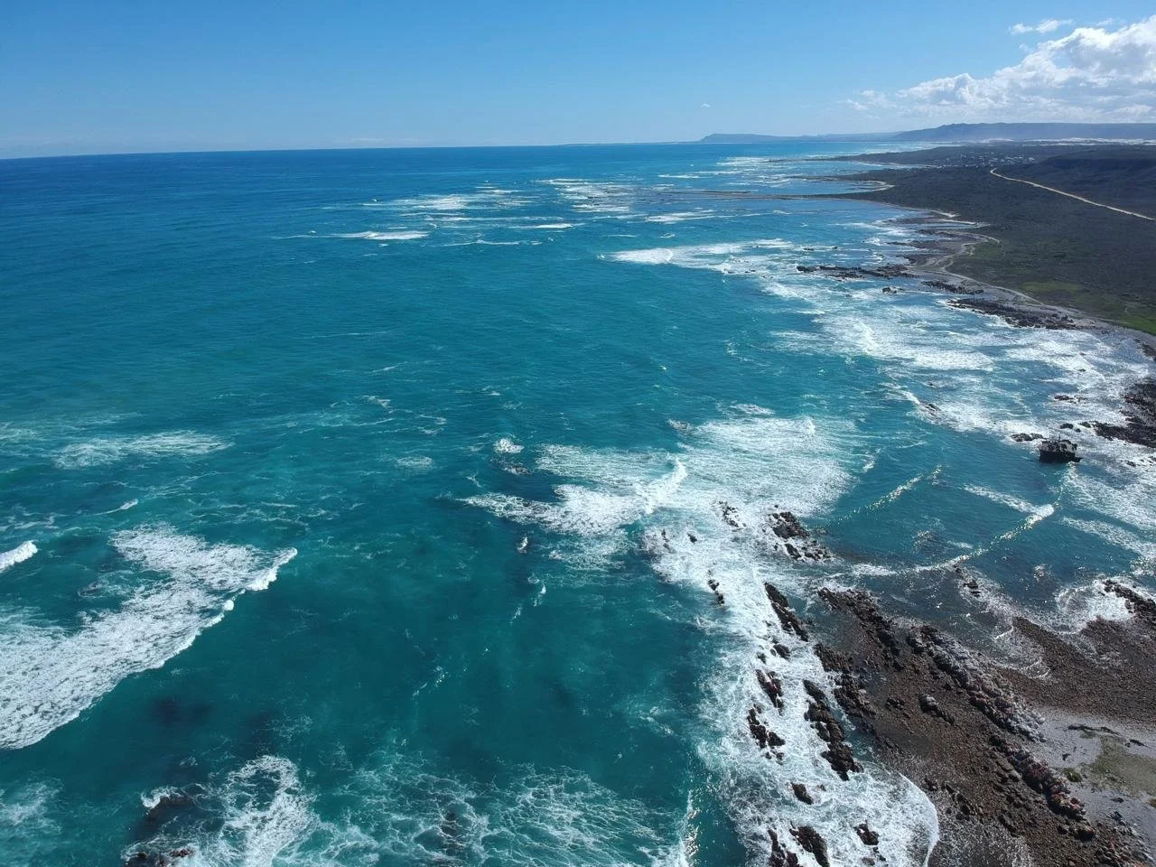 Aerial view of the ocean and shoreline with waves crashing onto rocks and a beach, with land and hills in the distance under a partly cloudy sky.