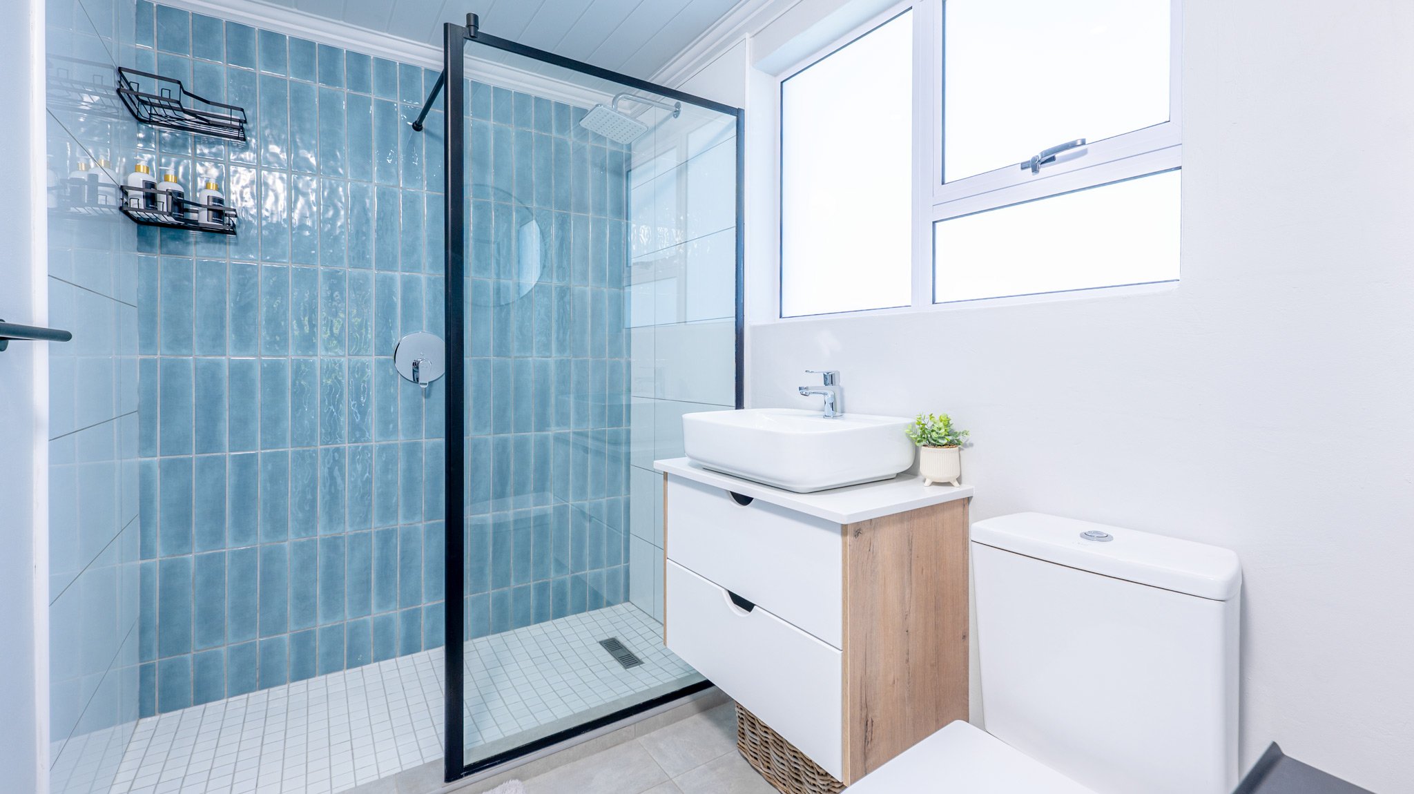 Modern bathroom with glass-enclosed shower, white sink on wooden vanity, toilet, and a window letting in natural light.