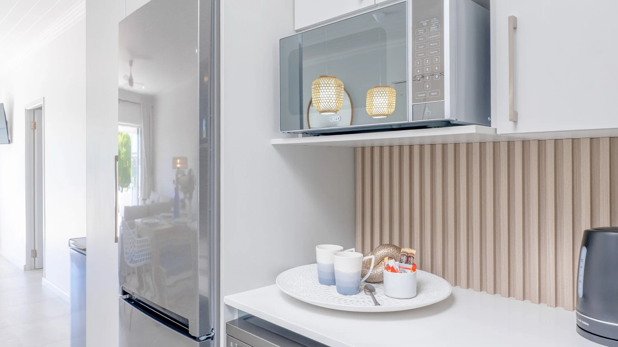 Kitchen with a stainless steel refrigerator, microwave, white countertop with a tray holding two cups, a small white jar with condiments, and a spoon, beige wood panel backsplash, and a black kettle.