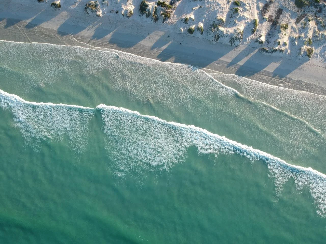 Aerial view of a sandy beach with ocean waves crashing onto shore, with rocky cliffs along the coastline.
