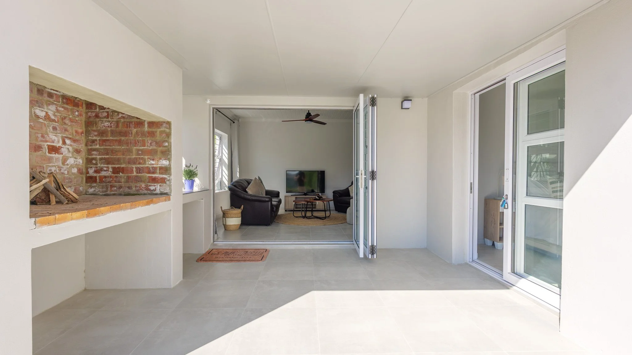 Patio area with open sliding glass doors leading to a living room with black leather furniture and a TV, a brick fireplace, a potted plant, and a beige tiled floor.
