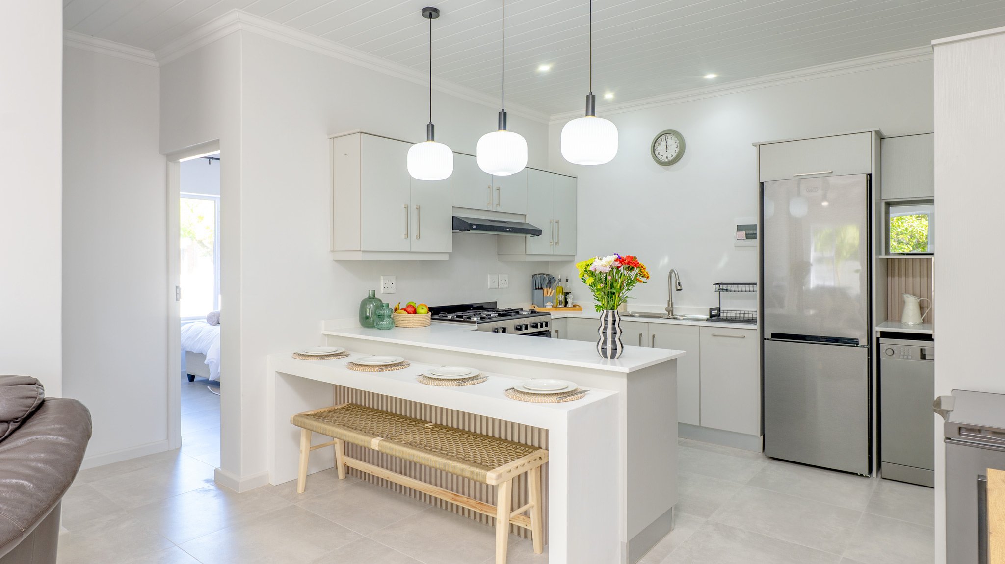 Modern kitchen with white cabinets, stainless steel refrigerator, and a kitchen island with a vase of flowers on top. Pendant lights hang above the island, and a clock is on the wall.
