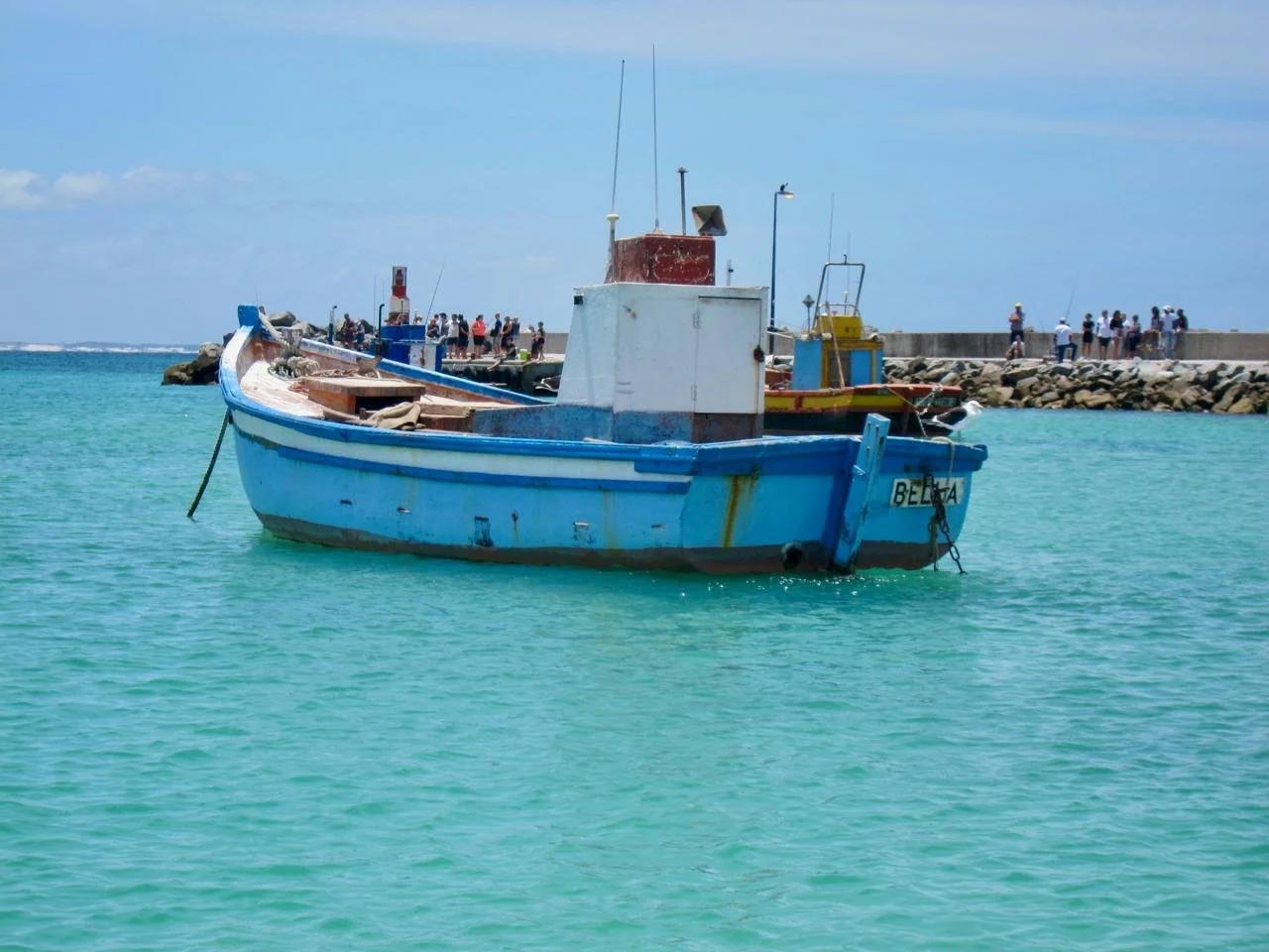 A blue boat floating in turquoise water with a concrete pier and people in the background.