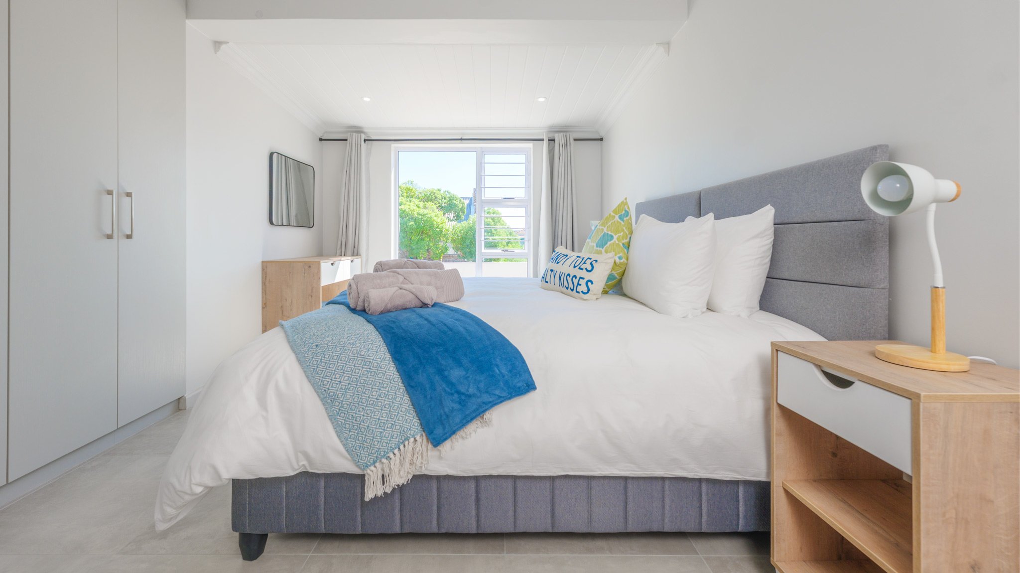 A bright bedroom with a large window, white walls, and a cozy bed with white linens, colorful pillows, and a blue throw blanket. There is a bedside table with a modern lamp and an outdoor view of trees.