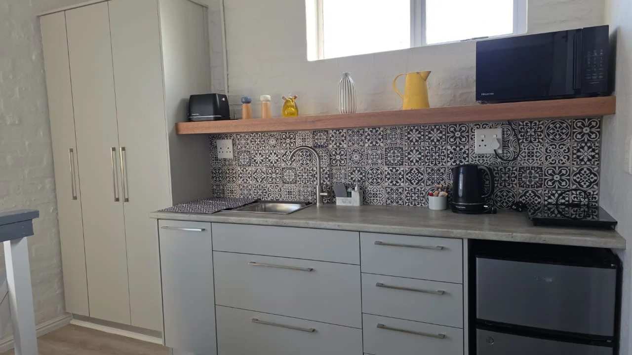 Modern kitchen with white cabinets, a concrete countertop, patterned black and white tile backsplash, and a wooden shelf above holding kitchen appliances and decorative items.
