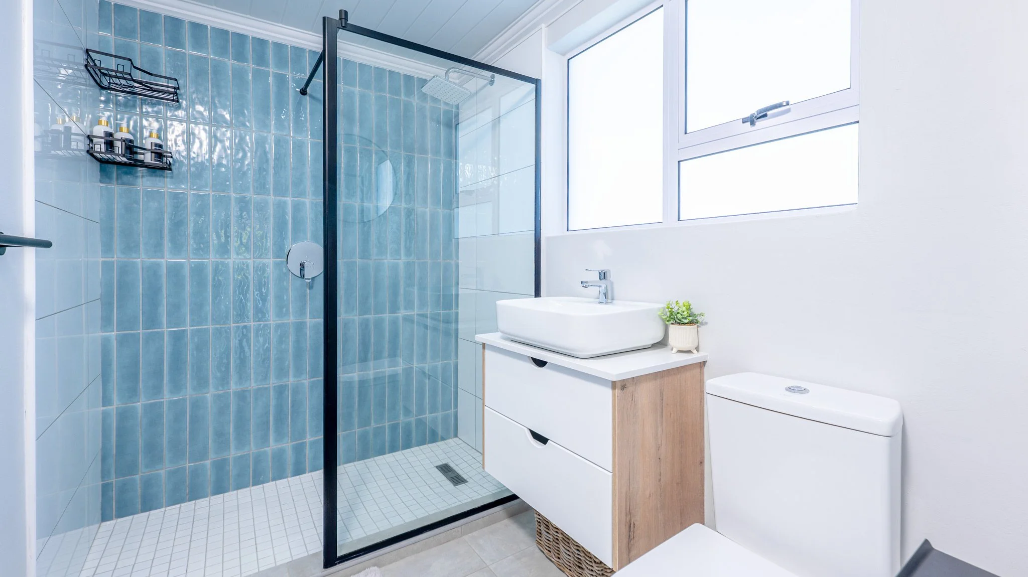 A modern bathroom featuring a walk-in shower with blue tiles, a white vanity with a vessel sink, a toilet, and a large window letting in natural light.