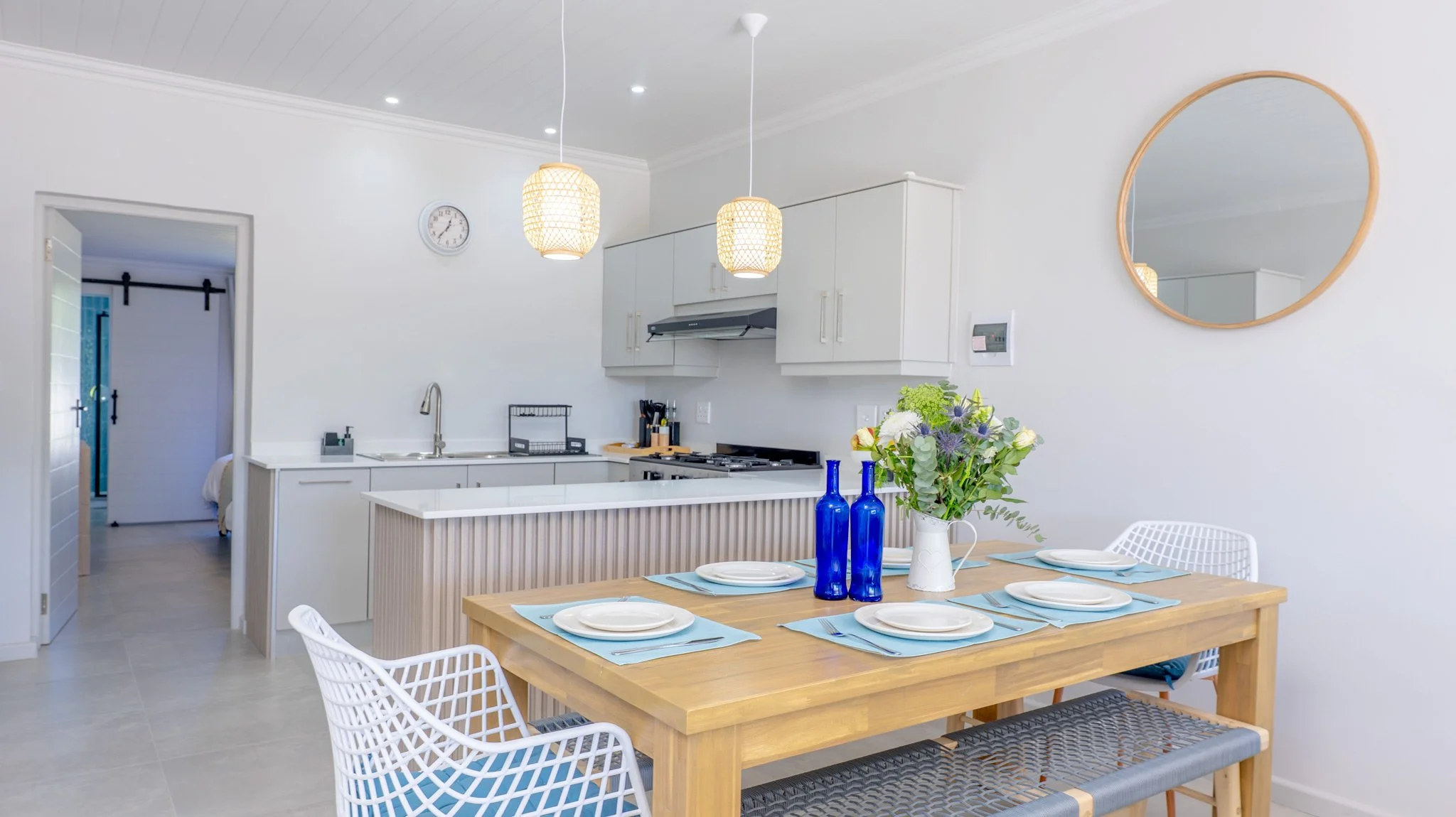 Dining area with a wooden table set with six plates and light blue placemats, with a white vase of flowers and blue bottles as a centerpiece, and white chairs. Kitchen in the background with white cabinets, a stove, and hanging pendant lights.