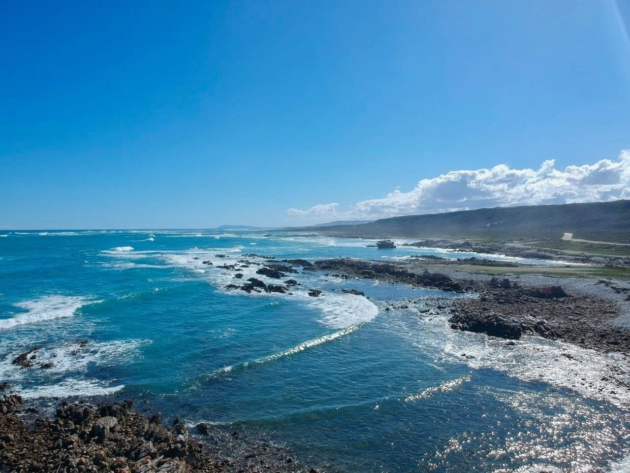 Scenic view of a rocky coastline with turquoise ocean waves under a bright blue sky with some clouds.
