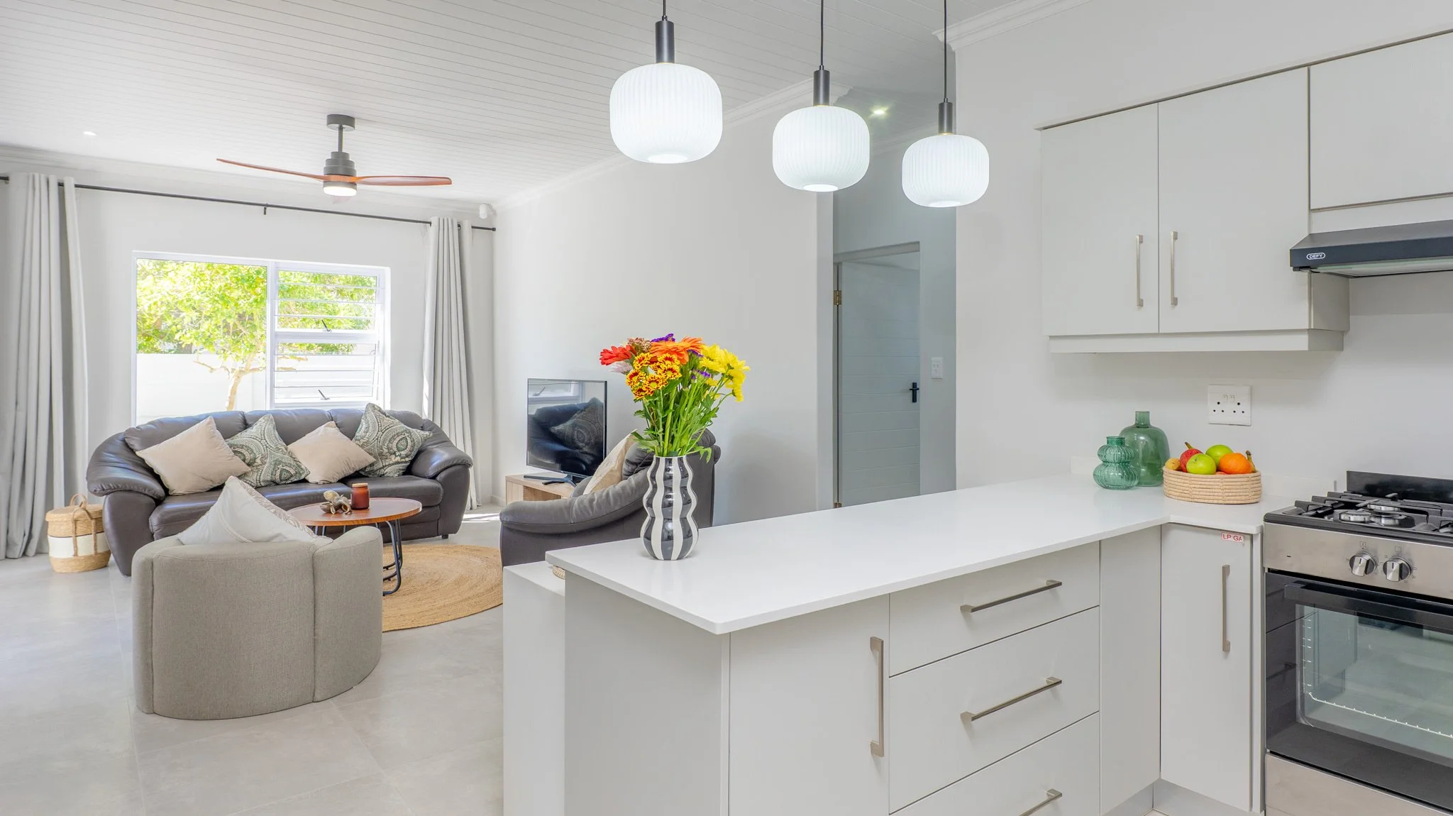 Living room with a brown sofa, beige armchair, and a small table, adjacent to a kitchen with a white countertop, decorative vases, and a fruit basket, illuminated by white hanging lights.