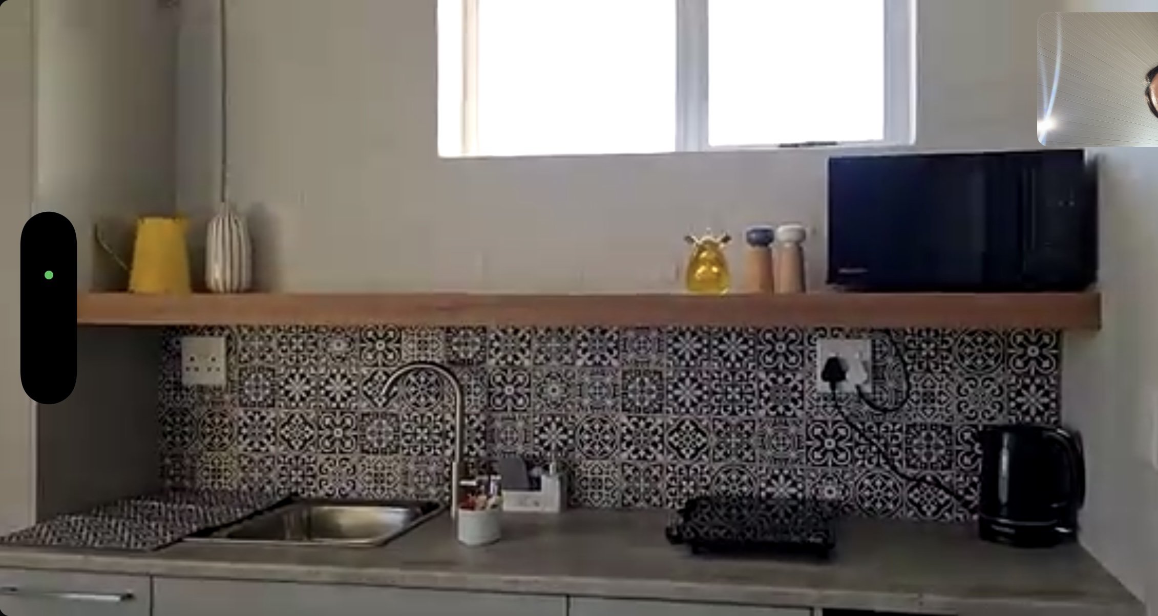 A kitchen counter with a patterned backsplash, a small sink, a dish soap dispenser, a kettle, a black bag, a microwave, and decorative jars, with a window above providing natural light.