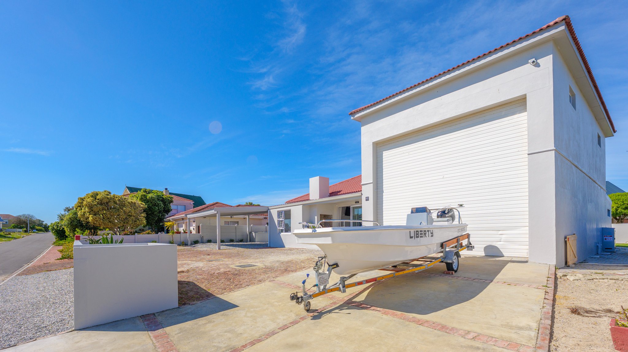 White house with a large garage door, a boat labeled 'Liberty' on a trailer in front, and a clear blue sky.