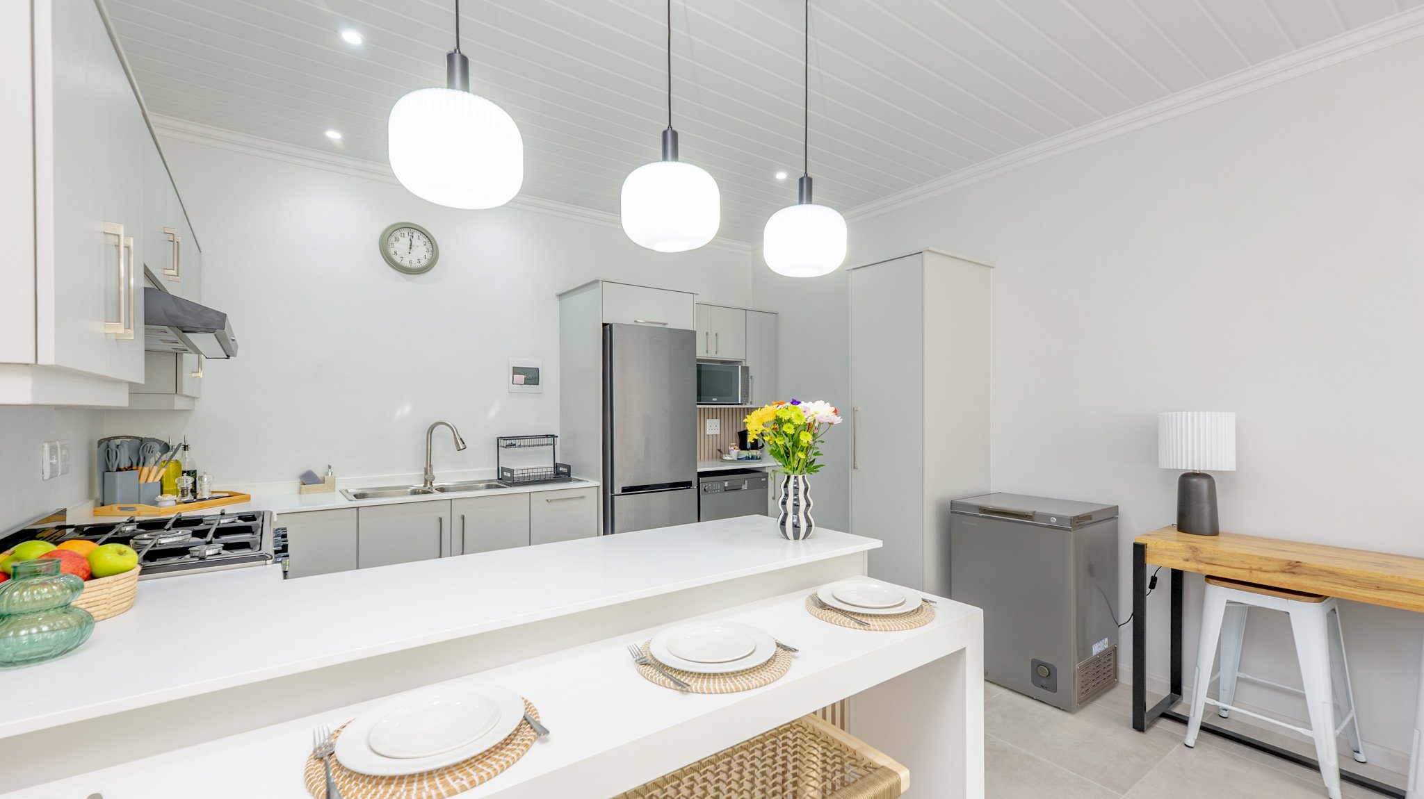 A modern kitchen with white cabinets, stainless steel appliances including refrigerator and microwave, a white island with place settings, and a wall clock. A vase with flowers is on the island, and there is a small wooden desk with a lamp and a stoo