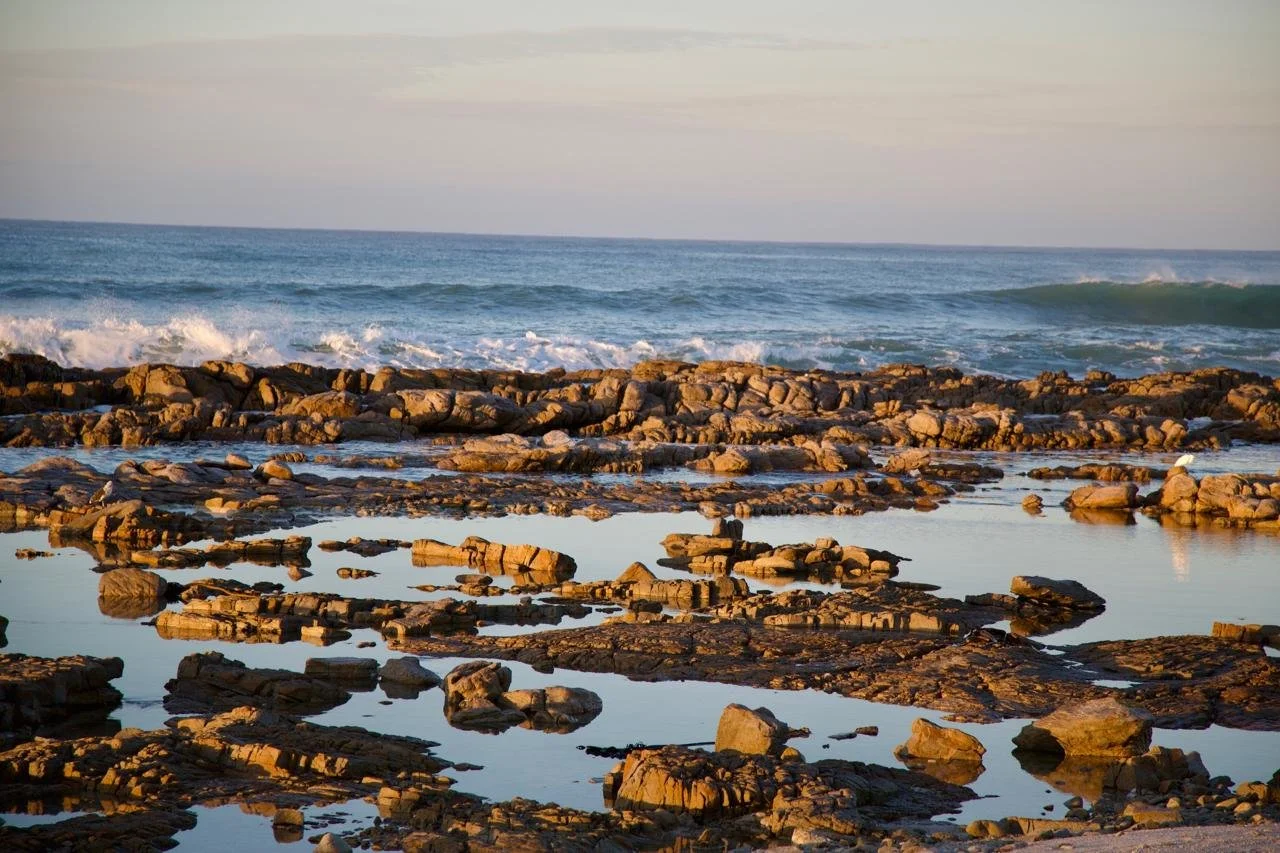 Scenic view of a rocky shoreline with water pools, waves, and the ocean in the background during sunset.