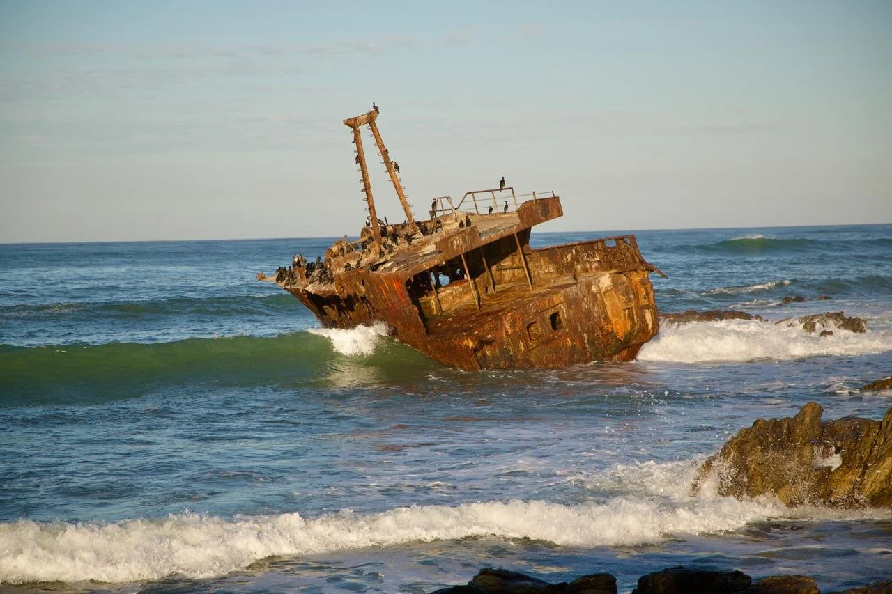 A rusted, abandoned shipwreck partially submerged in the ocean near a rocky shoreline, with small waves and a clear sky.