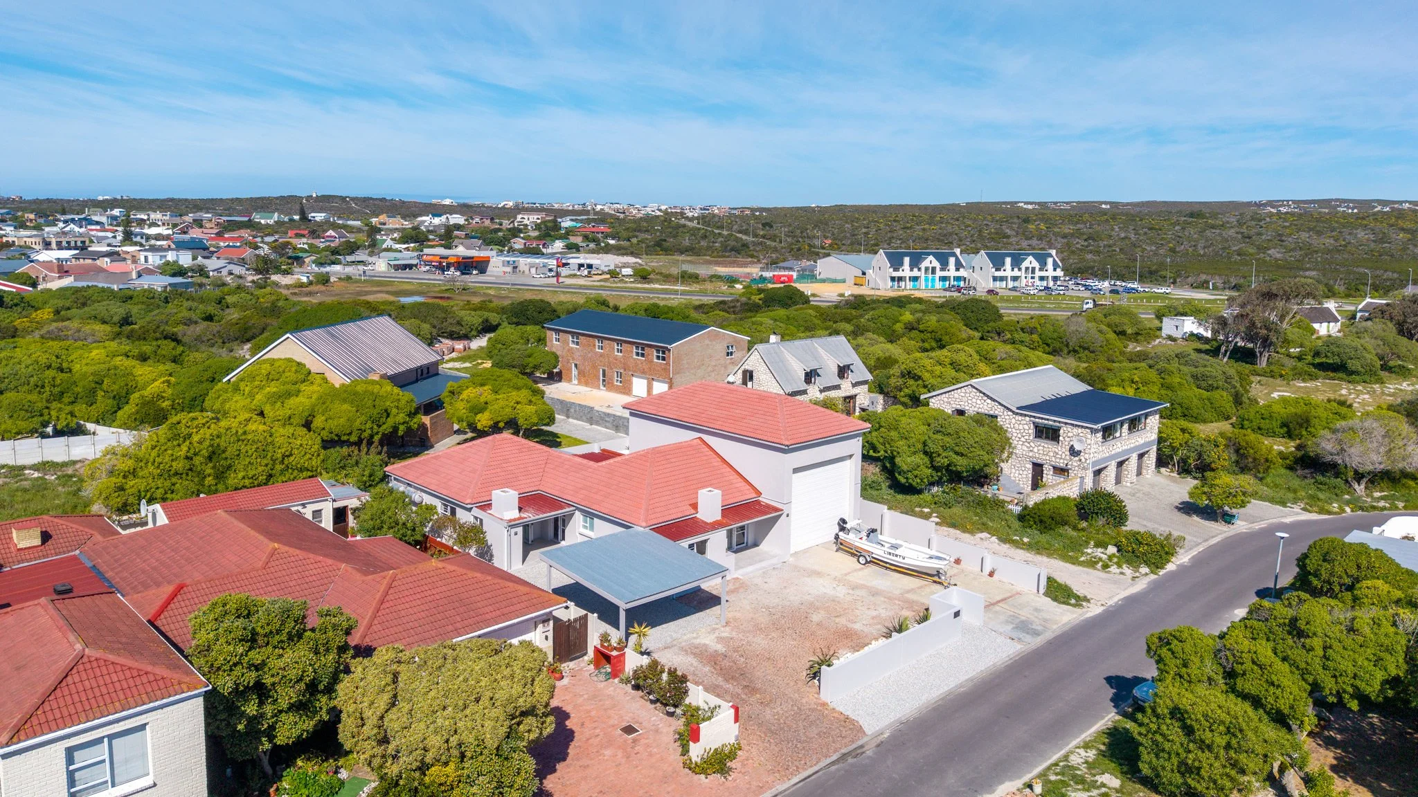 Aerial view of a suburban neighborhood near the coast with multiple houses, trees, and a distant view of a commercial area, under a blue sky.