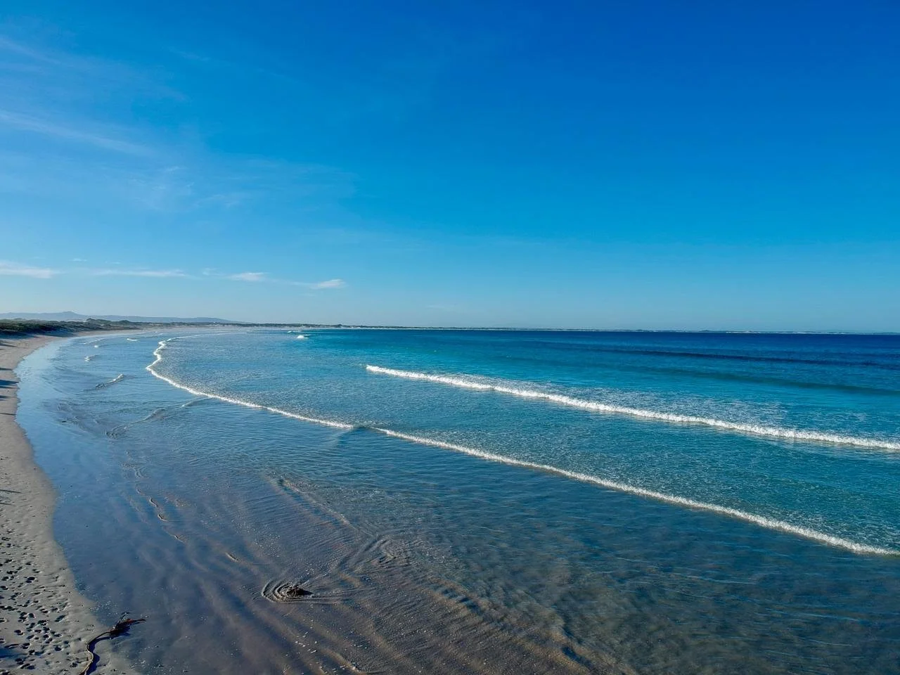 Calm ocean with small waves hitting sandy beach under a clear blue sky.