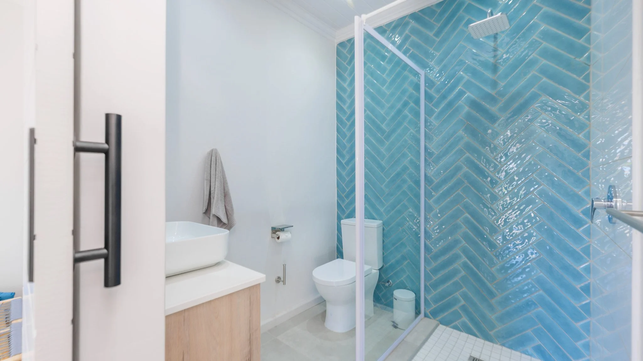 Modern bathroom with a glass-enclosed shower area featuring blue herringbone tiles, a white toilet, a white sink with a wooden cabinet, a towel hanging on the wall, and a small trash bin.