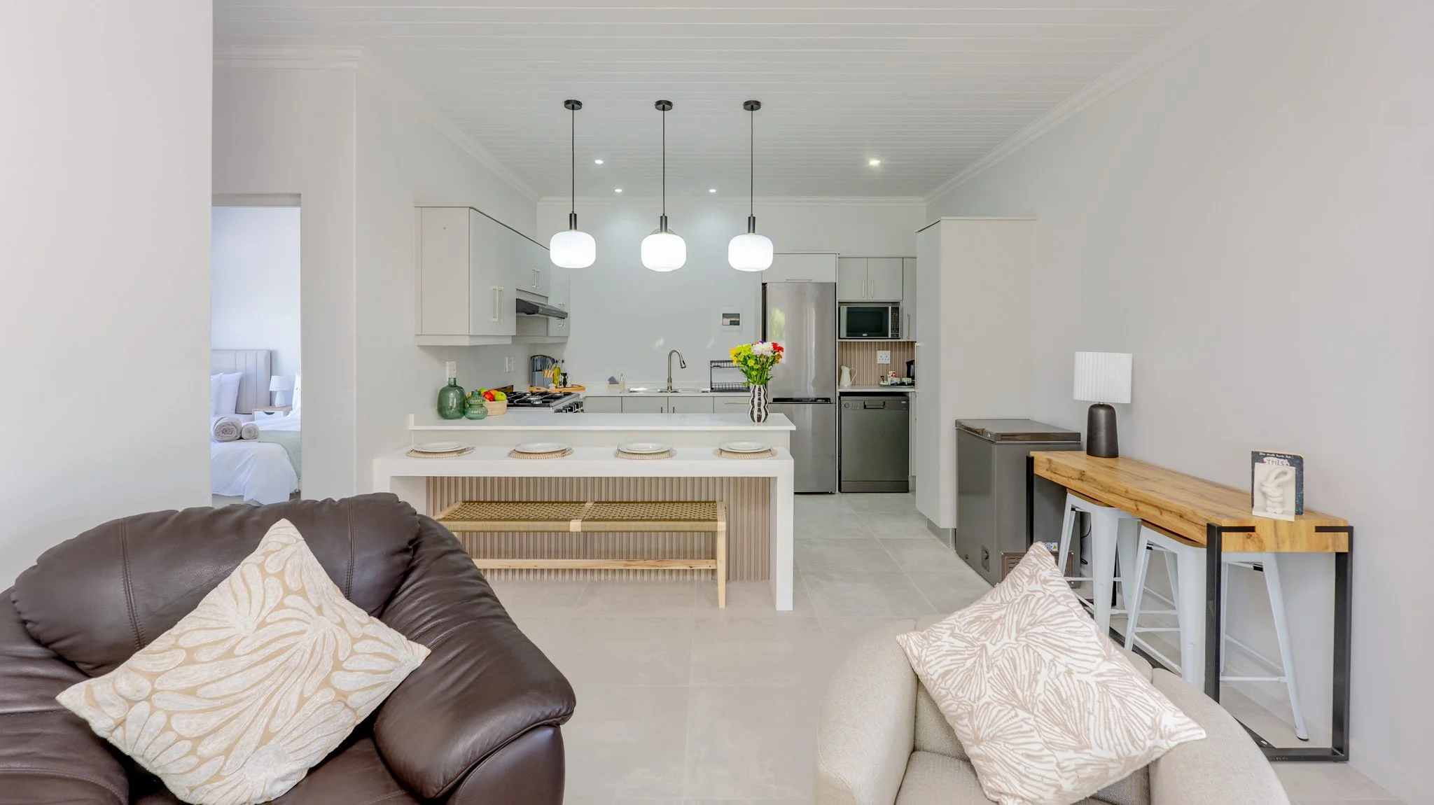 Living room with brown sofa, beige armchair, decorative pillows, open kitchen with white cabinetry, island with table setting, pendant lights, and bar stools, doorway leading to bedroom with bed and nightstand.