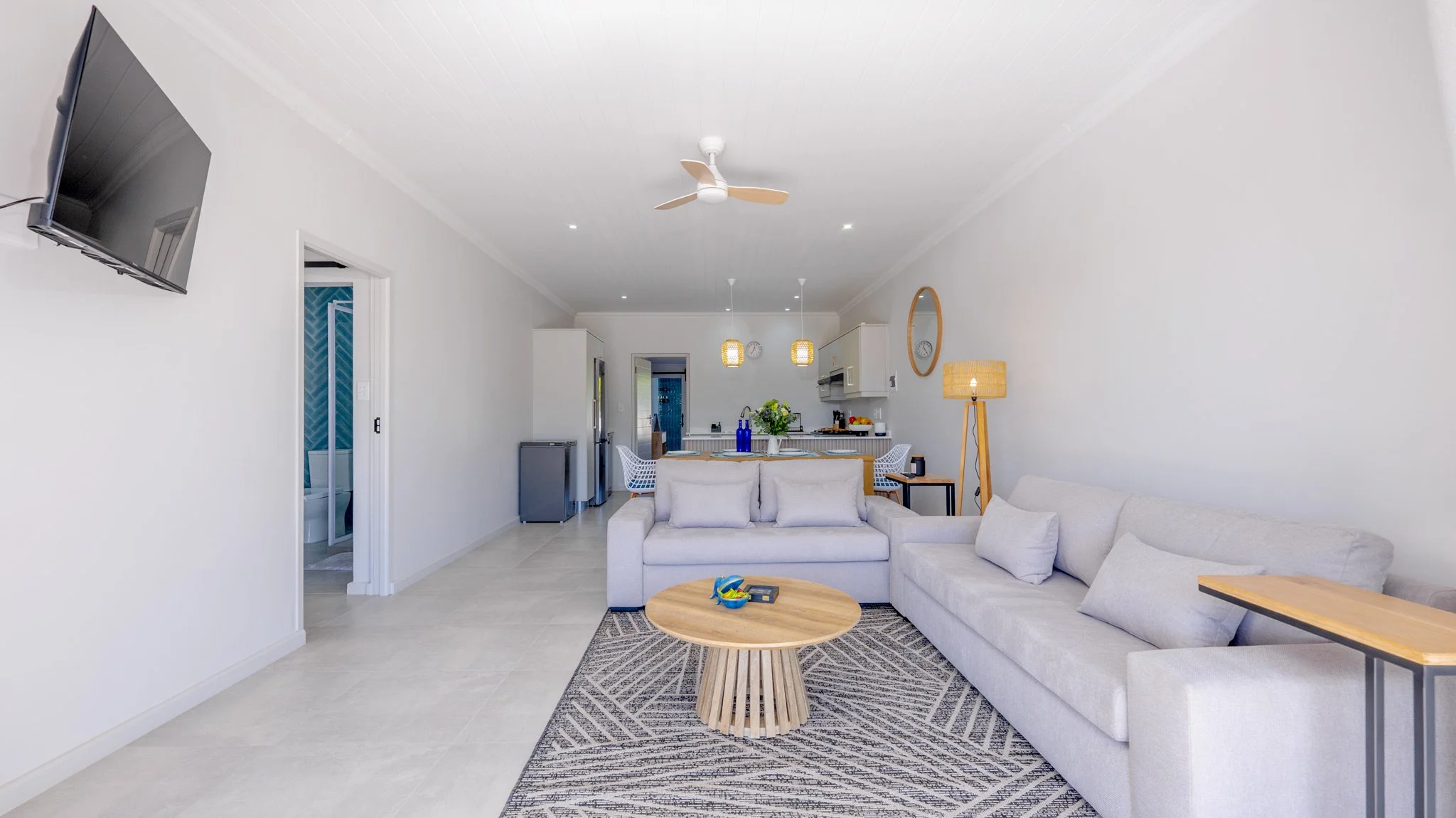 Living room with white walls, light-colored sofa, black and wood side table, round wooden coffee table, ceiling fan, flat-screen TV, rug, and a floor lamp in a modern open-concept space with a kitchen in the background.