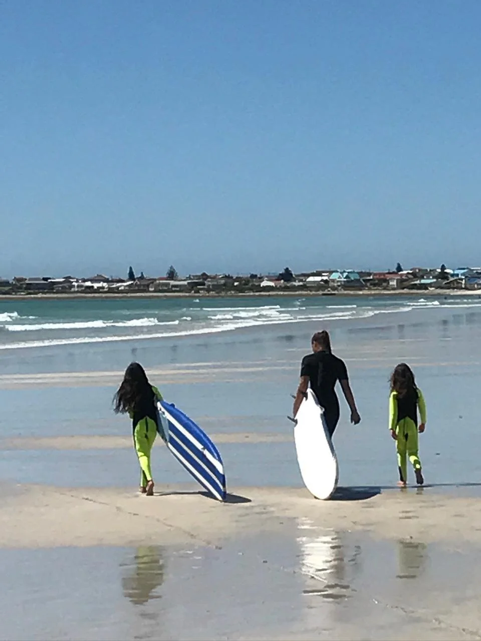 Three people walking on the beach with surfboards, two young girls in bright yellow wetsuits and a woman in black, carrying surfboards, near the shoreline with houses in the background.