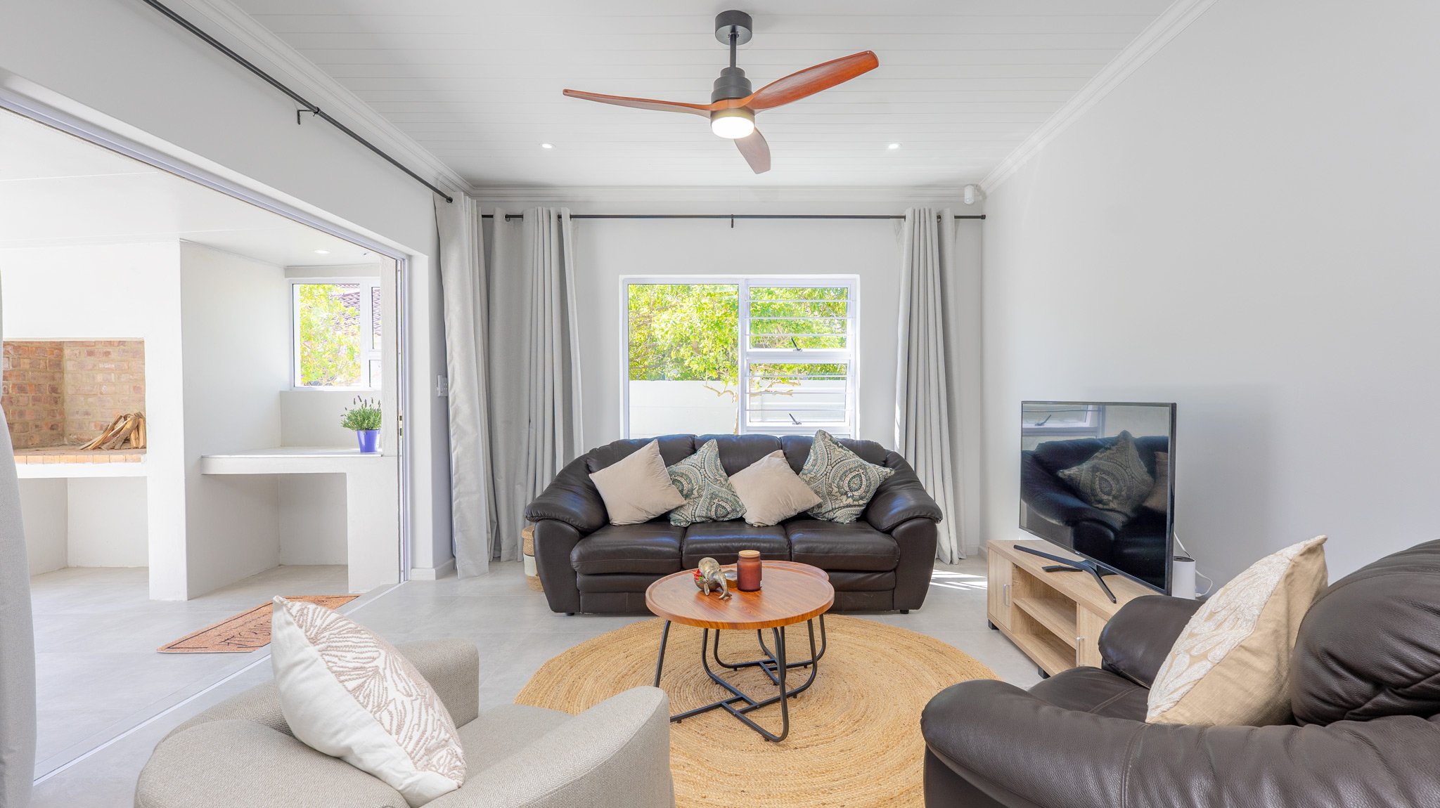 Bright living room with white walls, a window with trees outside, white curtains, and a ceiling fan. Contains a brown leather sofa with pillows, a beige armchair with pillows, a small round wooden coffee table with decorative items, a flat-screen TV 