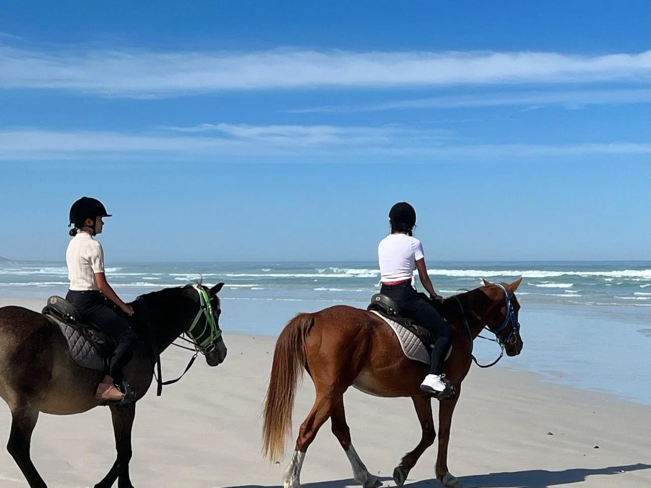 Two women riding horses on a beach with the ocean and blue sky in the background.