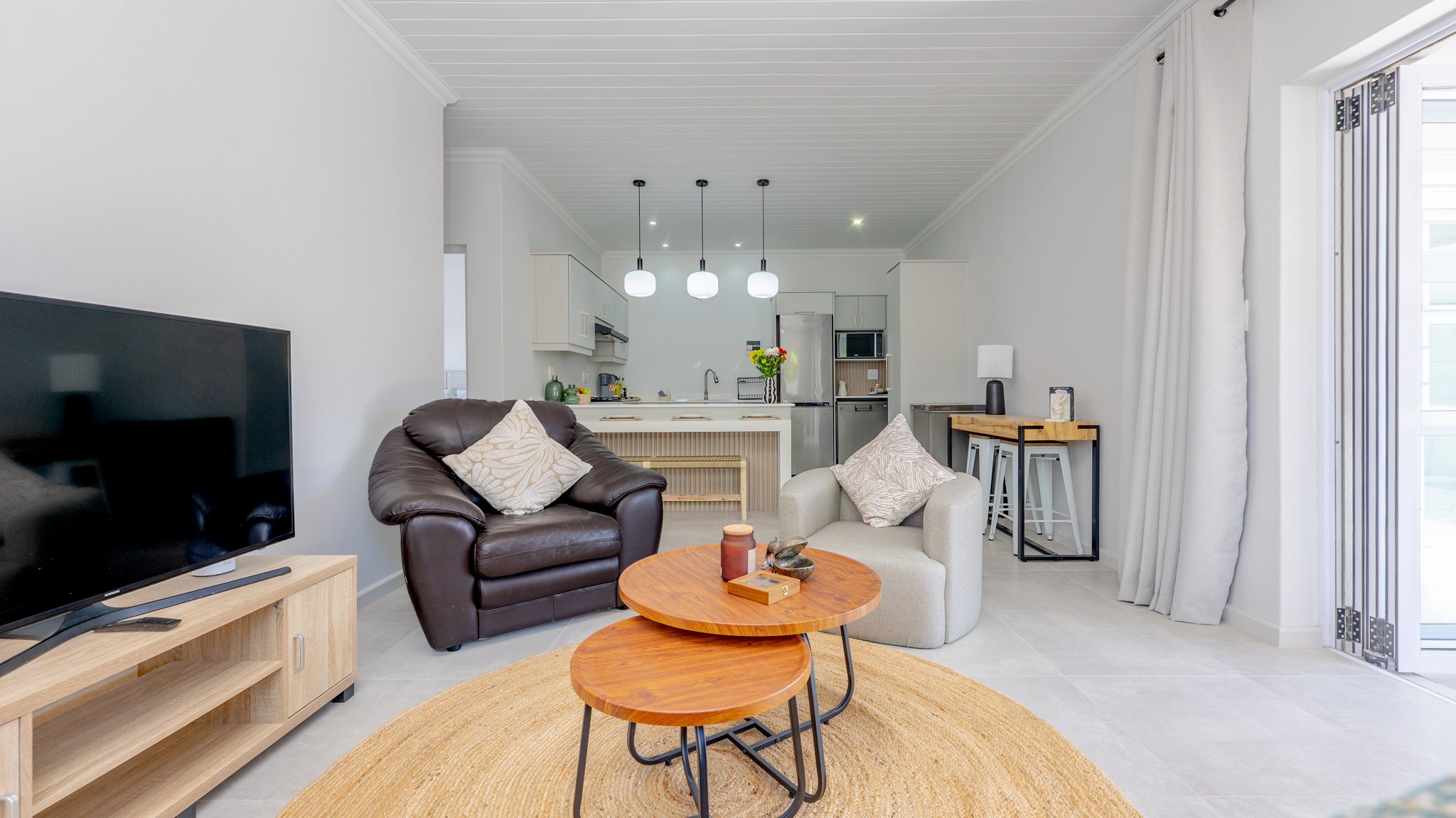 Living room with a black leather armchair, a beige armchair, a wooden coffee table, a TV on a wooden stand, and a round rug. In the background, a kitchen with white cabinets, a refrigerator, and pendant lighting. Large window with curtains on the rig