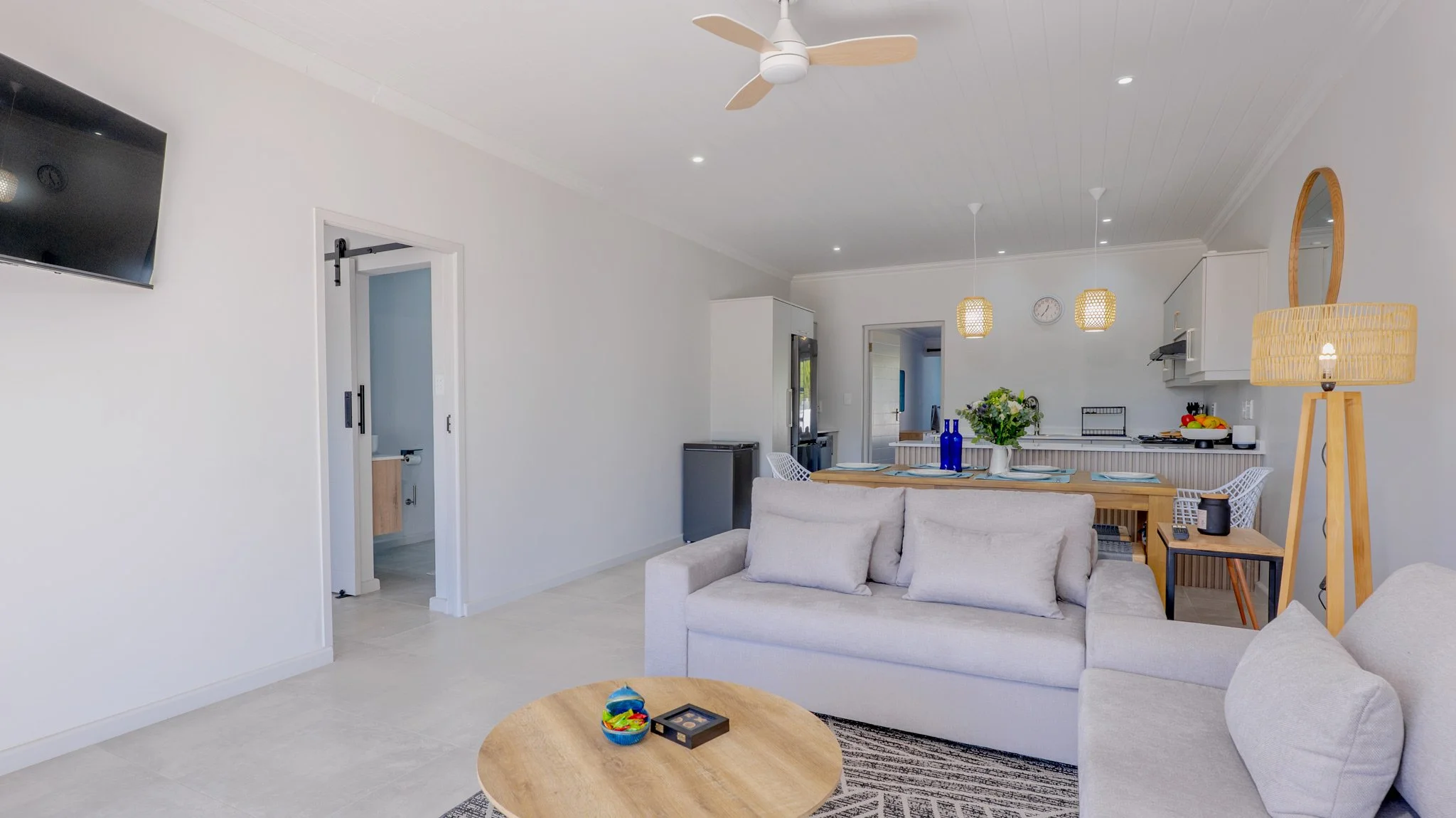 Bright open living room with white walls, beige sofas, wooden coffee table, and modern decor. In the background, a dining area and kitchen with pendant lights, a clock, and a vase with flowers on the table.