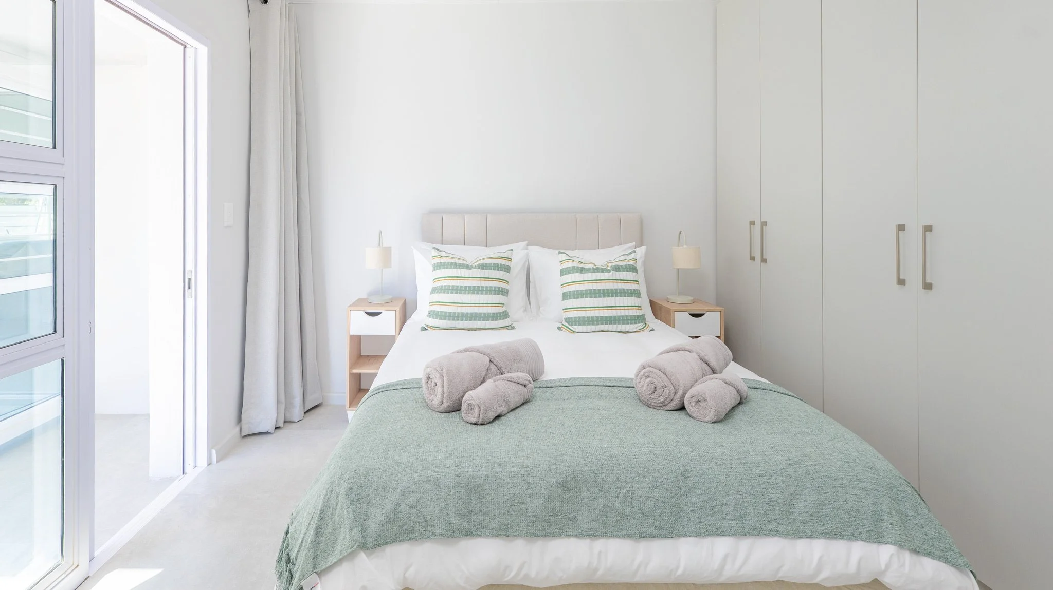 A neatly made bed with a green blanket, white sheets, and two striped pillows in a bright, white bedroom. Folded towels are placed on the bed, and there are two small nightstands with lamps on either side of the bed. A large closet with white doors i