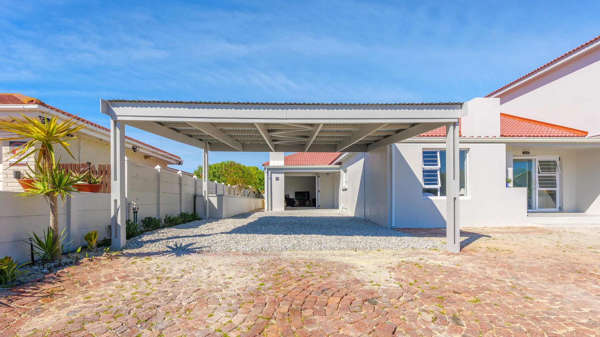 Front view of a house with a white exterior, red tiled roof, and a covered carport with gravel ground, adjacent to a brick-paved driveway, and clear blue sky.