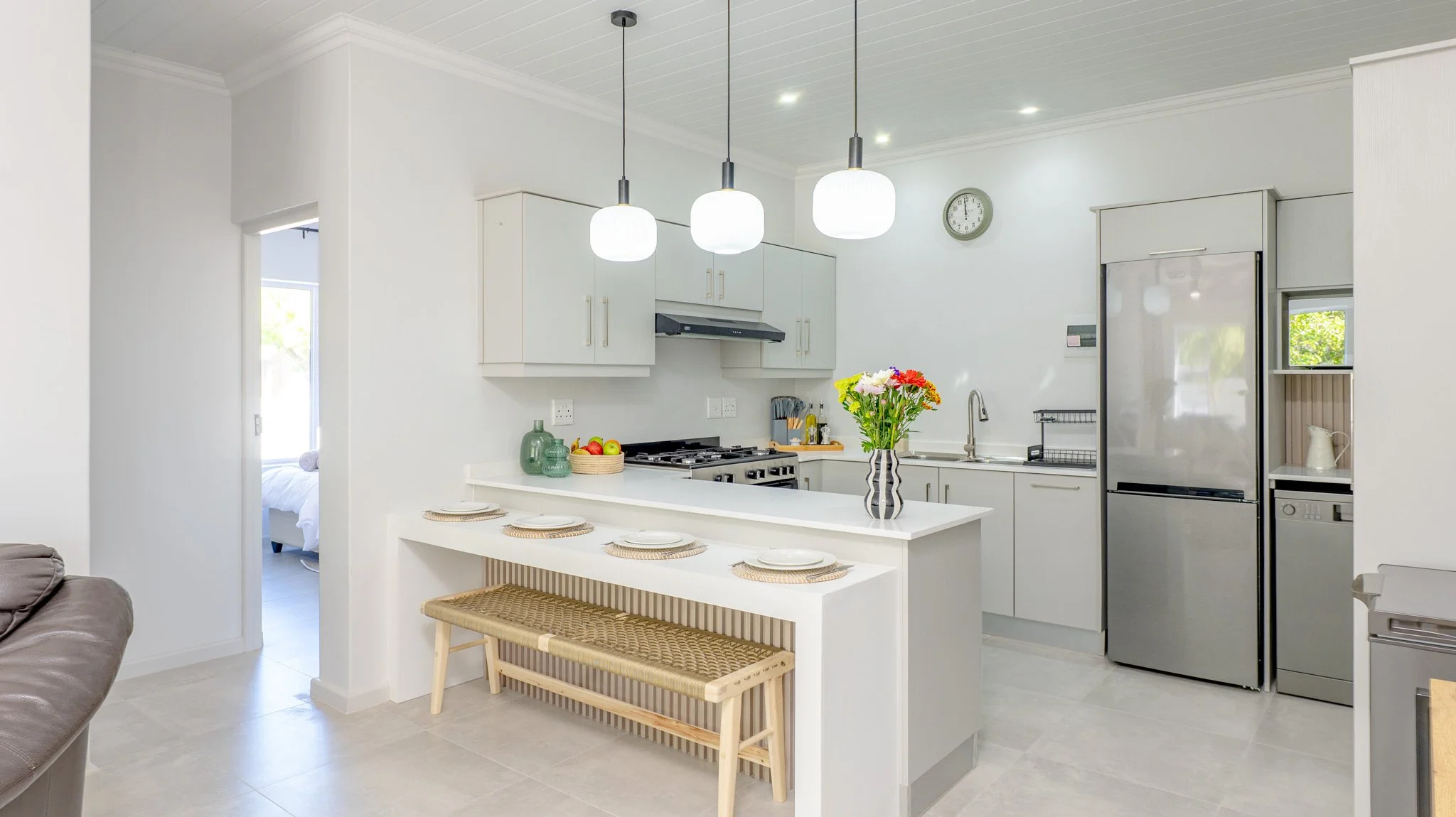 Modern kitchen with white cabinets, stainless steel refrigerator, black stove, and a white countertop island with a vase of colorful flowers, set against white walls and ceiling with three hanging pendant lights.