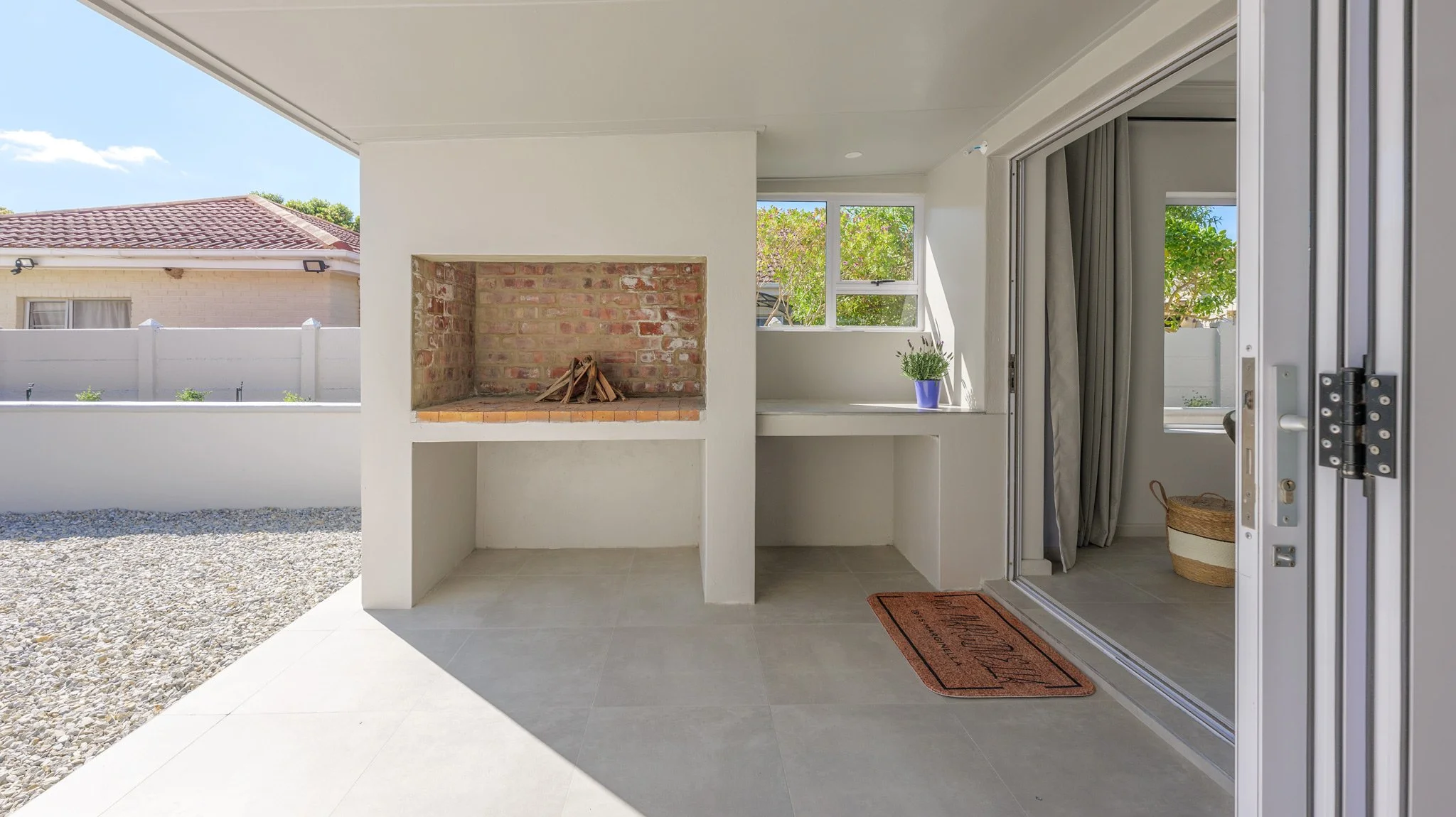 Covered patio area with built-in brick fireplace, side table with potted plant, sliding glass door with curtains, and gravel yard outside under sunny sky.